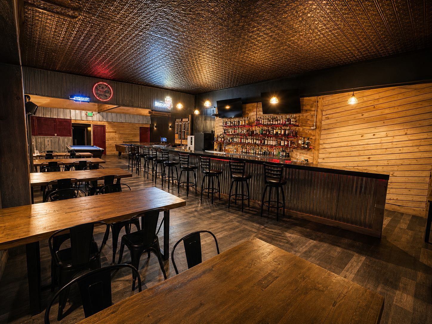 Interior of the historic 1904 bar with reclaimed wooden tables and black chairs, a long central saloon counter with stools, shelves with overflowing with bottles, TV screens, and neon signs, illuminated by small hanging pendant lights.