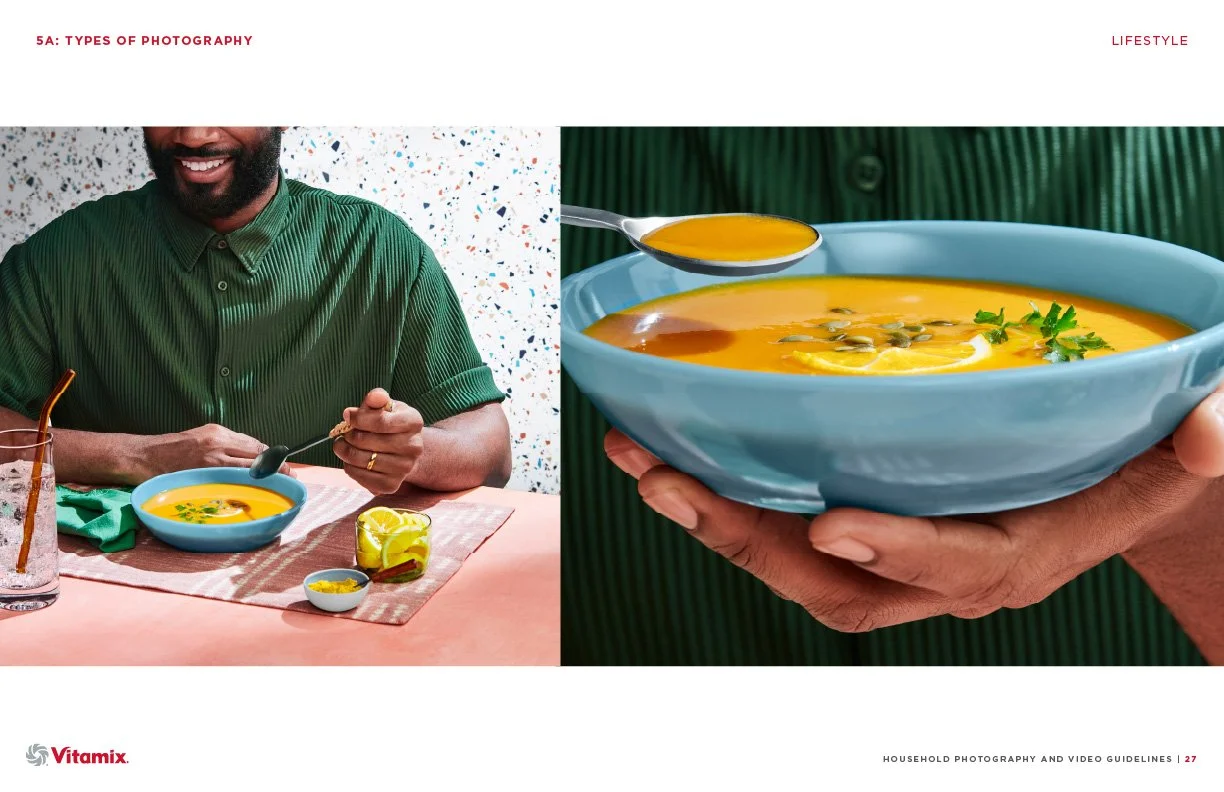 A person holding a blue bowl of orange-colored soup garnished with herbs and pumpkin seeds, with a spoonful of soup, a glass of water with a straw, lemon slices, and a small bowl of spices on a table.