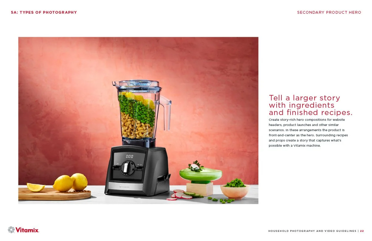 A Vitamix blender filled with colorful ingredients such as green peas, yellow lemon slices, and nuts. The blender is on a white surface with lemons on a cutting board to the left and fresh vegetables and herbs to the right, set against a pink texture