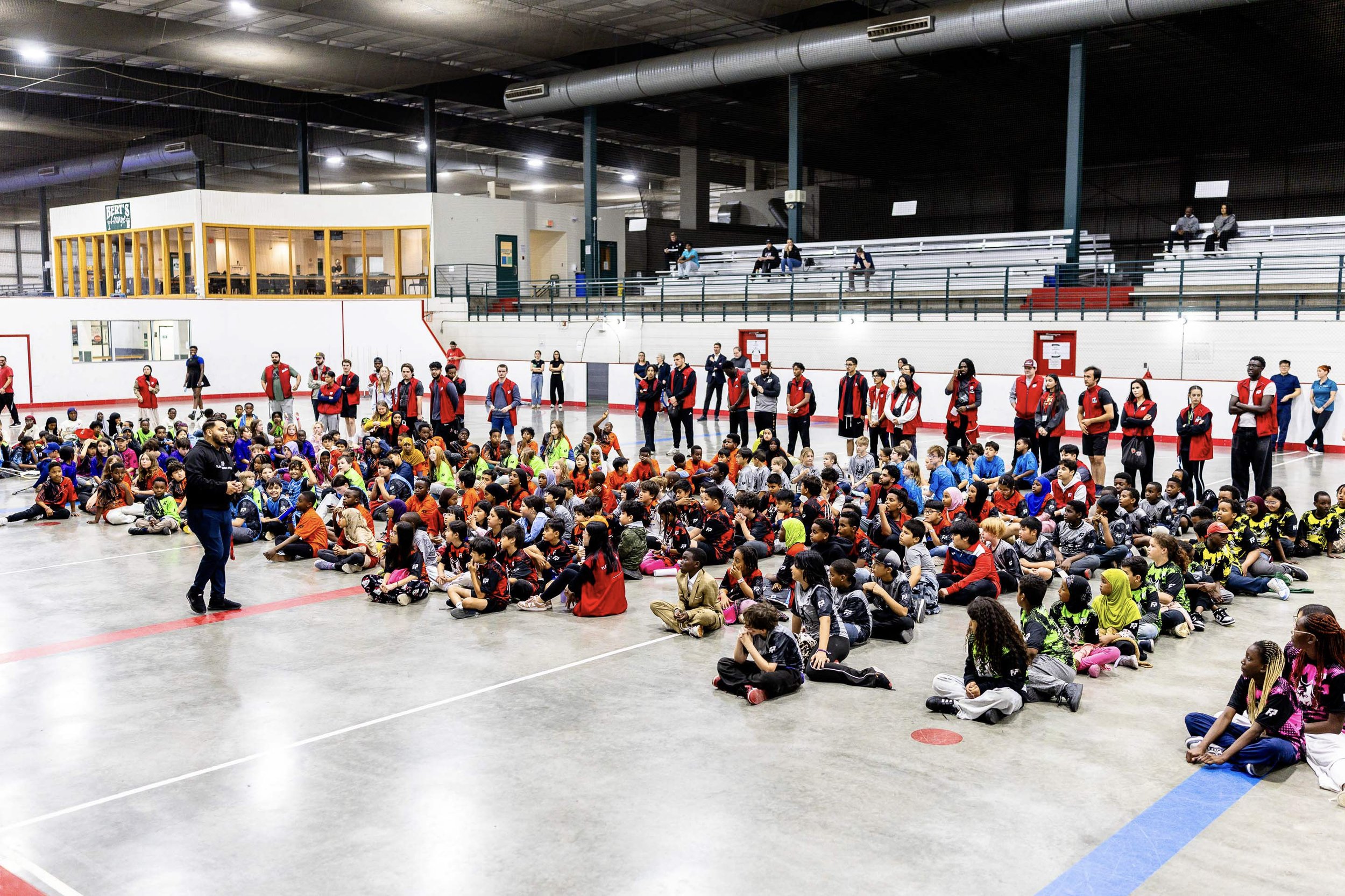 A large indoor gymnasium with a group of children sitting on the floor, listening to an adult speaker. Several adults are standing at the back, some wearing red jackets. Spectators are seated on bleachers in the background.
