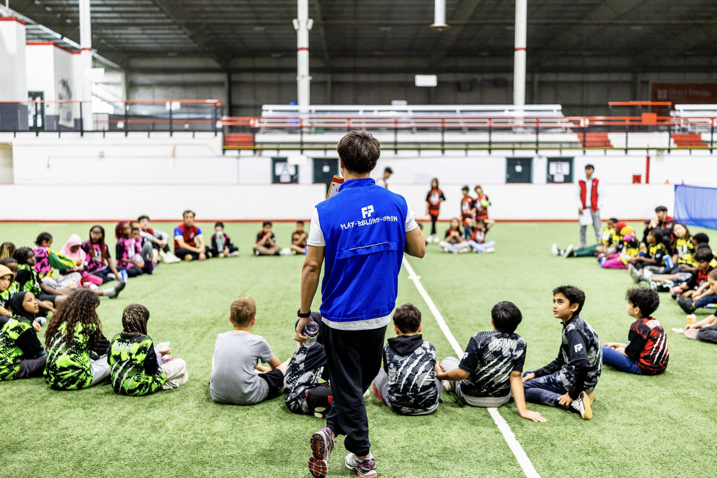 Children sitting on artificial turf inside a sports arena, listening to a coach in a blue vest with the words 'Play, Belong, Grow' written on the back, as part of a sports training session.