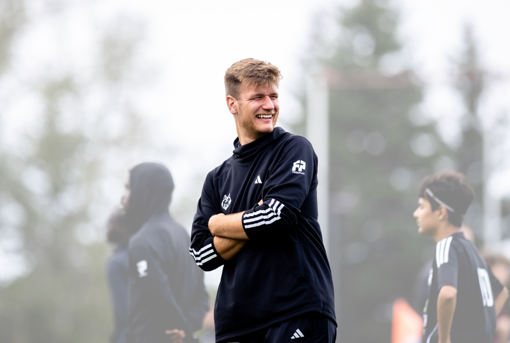 A young man with blond hair smiling and standing with arms crossed on a sports field, with other people in athletic clothing in the background, outdoors on a cloudy day.