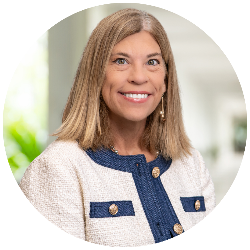 Portrait of a smiling woman with shoulder-length light brown hair, wearing a cream and navy blazer with gold buttons, and pearl earrings, standing in a bright, modern indoor setting.