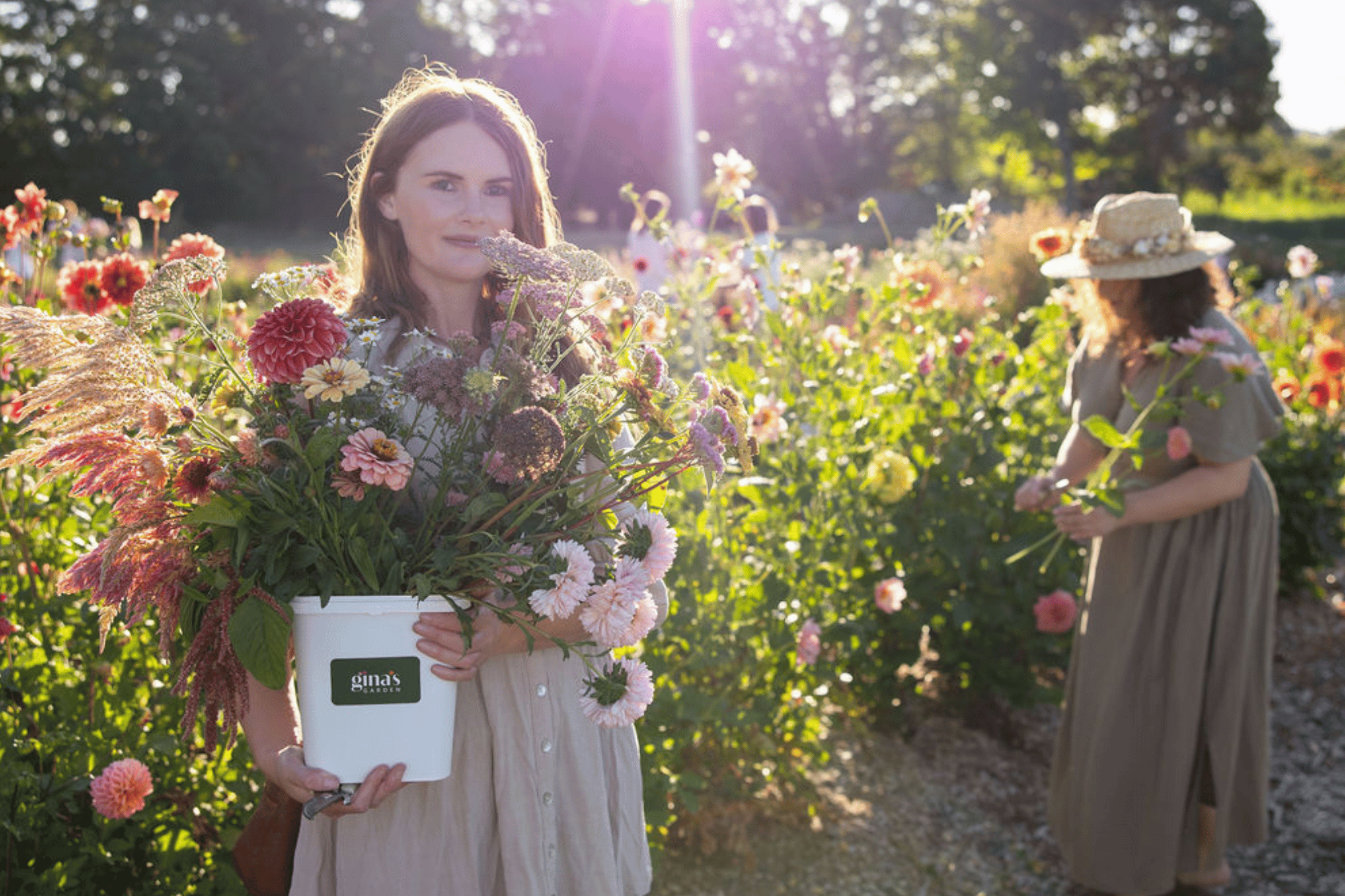 A woman holding a white bucket filled with pink, purple, and white flowers in a garden. Another woman in a large straw hat and gray dress is picking flowers in the background. The scene is lit by sunlight.