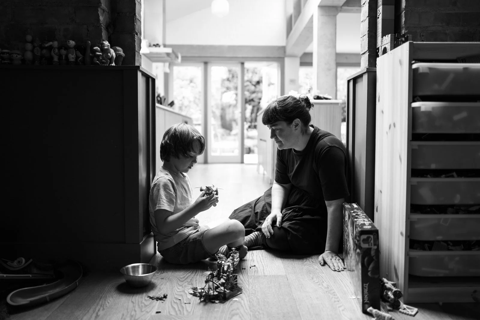 A woman and a young boy sit on the floor of a room, playing with building blocks and toys. The room has large windows in the background allowing natural light, and shelves with toys on the left.