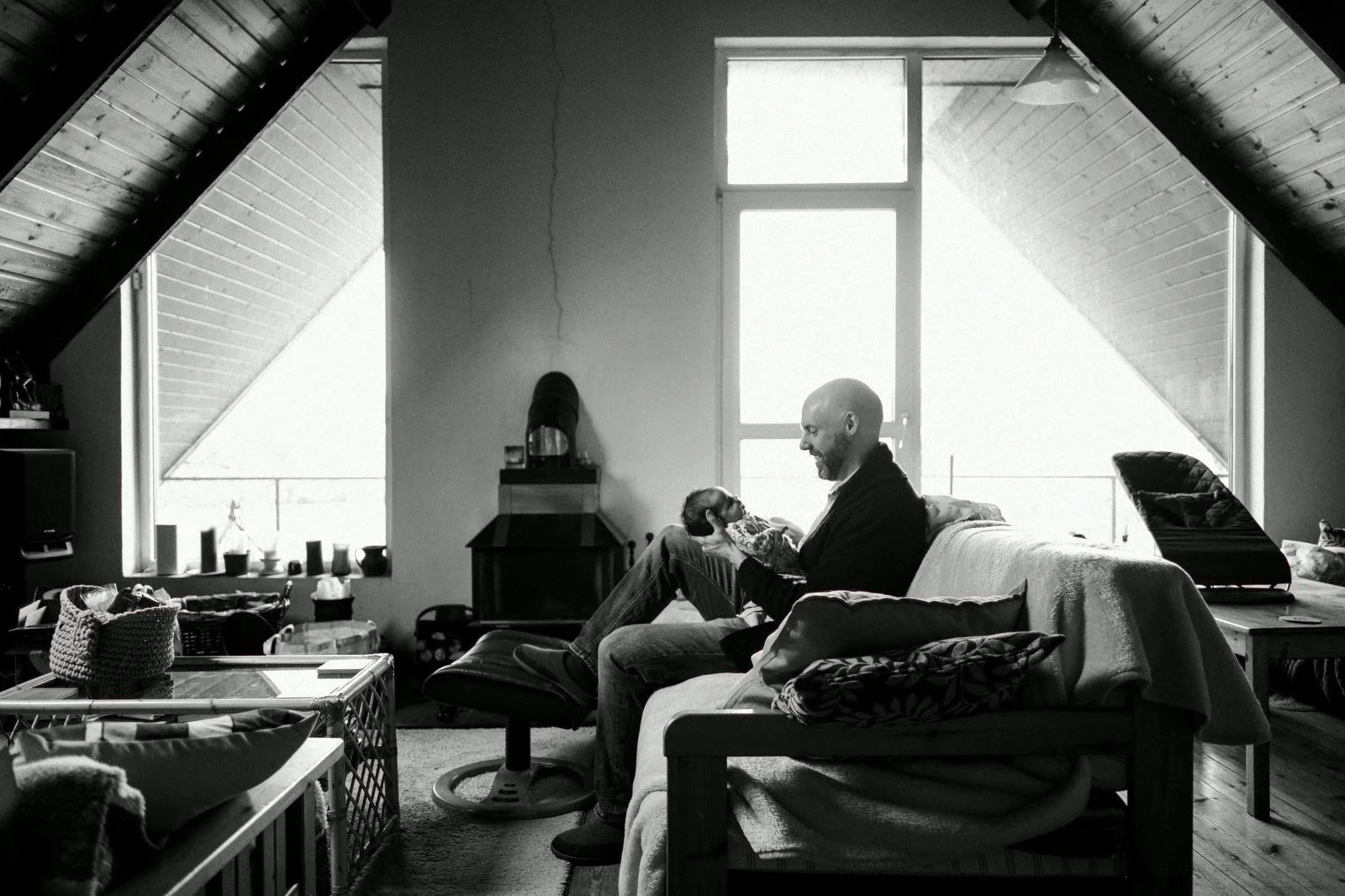 Man sitting on a couch, holding a small dog in his hands, smiling, in a cozy attic living room with large windows, sloped wooden ceiling, and various objects on tables and shelves.