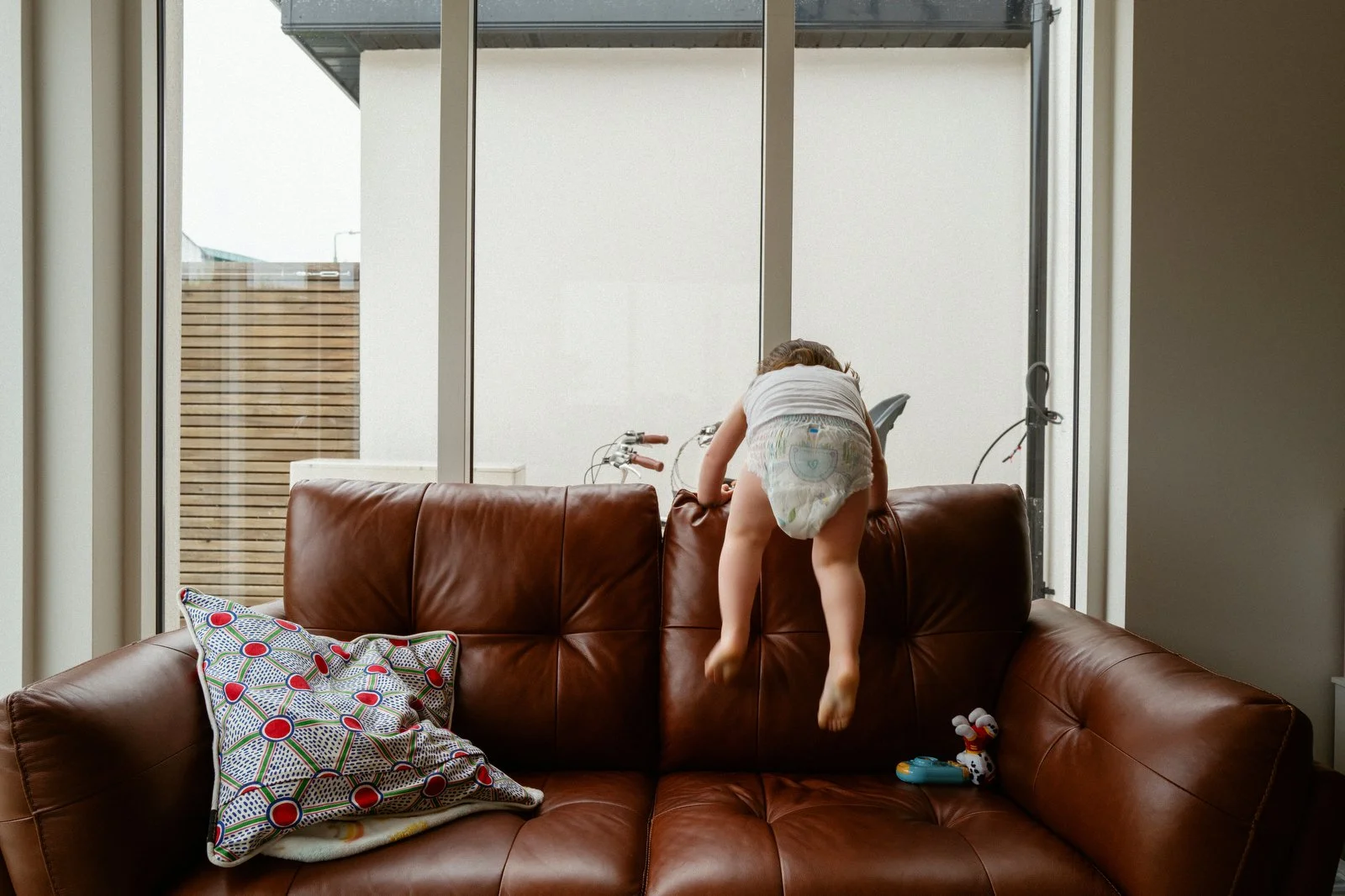A child in a diaper climbing on a brown leather couch inside a house near large windows with a backyard view.