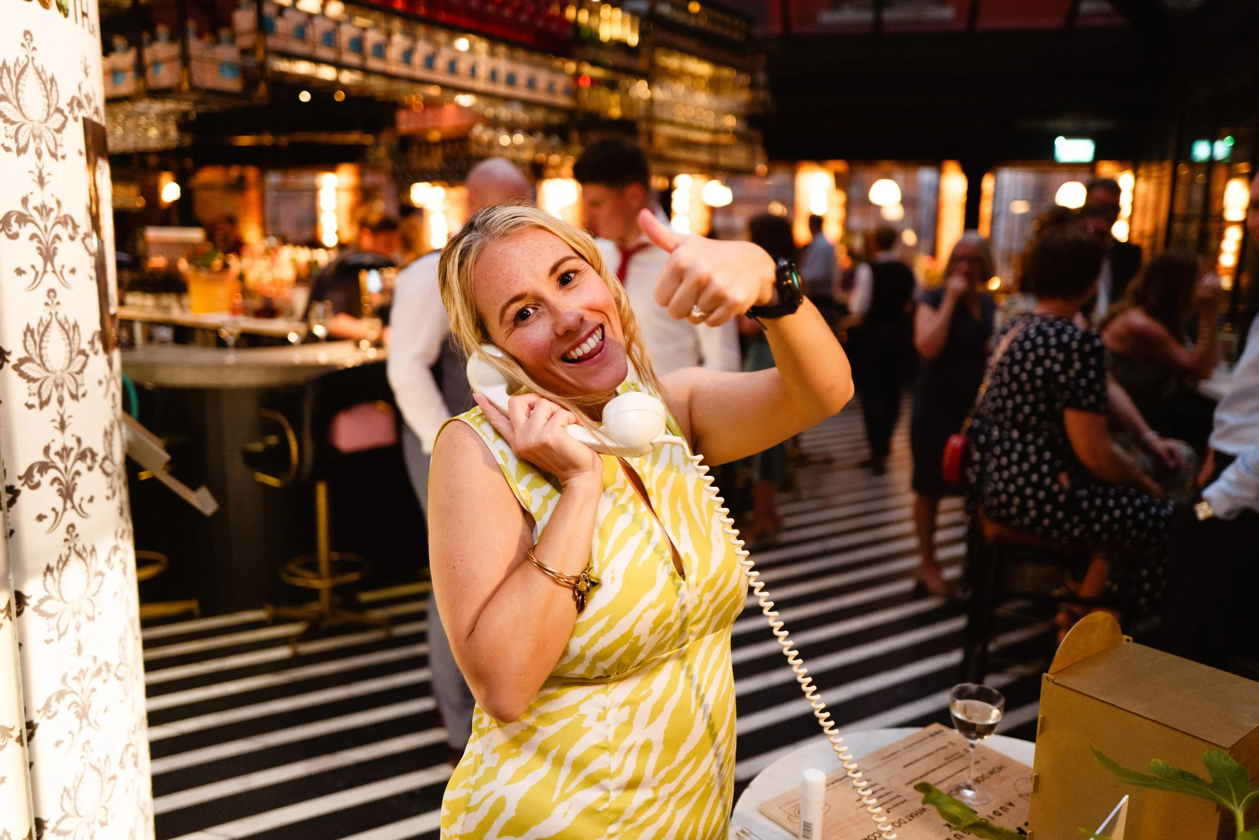 A woman in a yellow and white dress talking on a vintage telephone at a lively bar or restaurant, smiling and giving a thumbs up, with a busy background of people and bar activity.