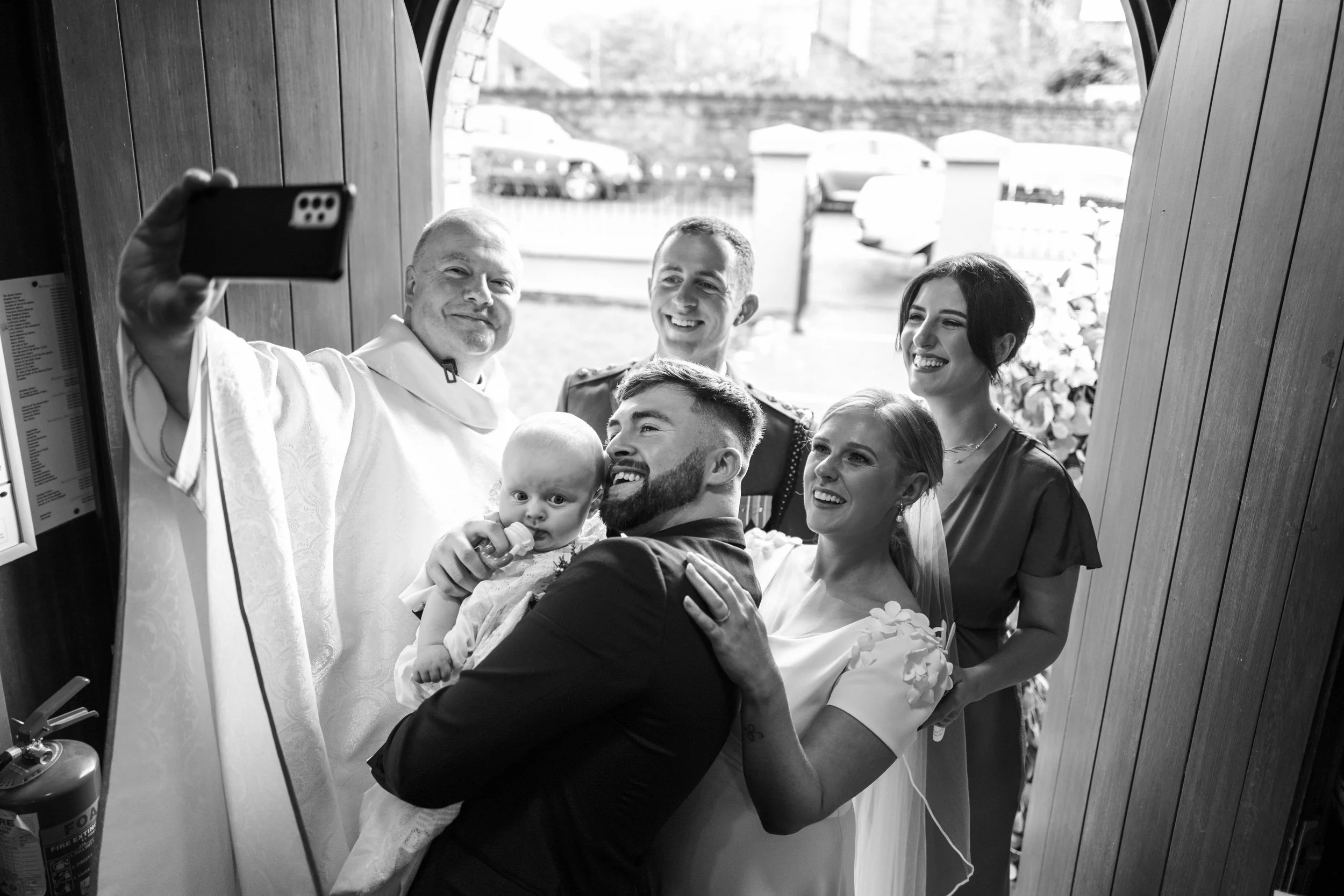 People taking a group selfie inside a small doorway. The group includes a man in a suit holding a baby, six others smiling, and a priest or minister in white robes.