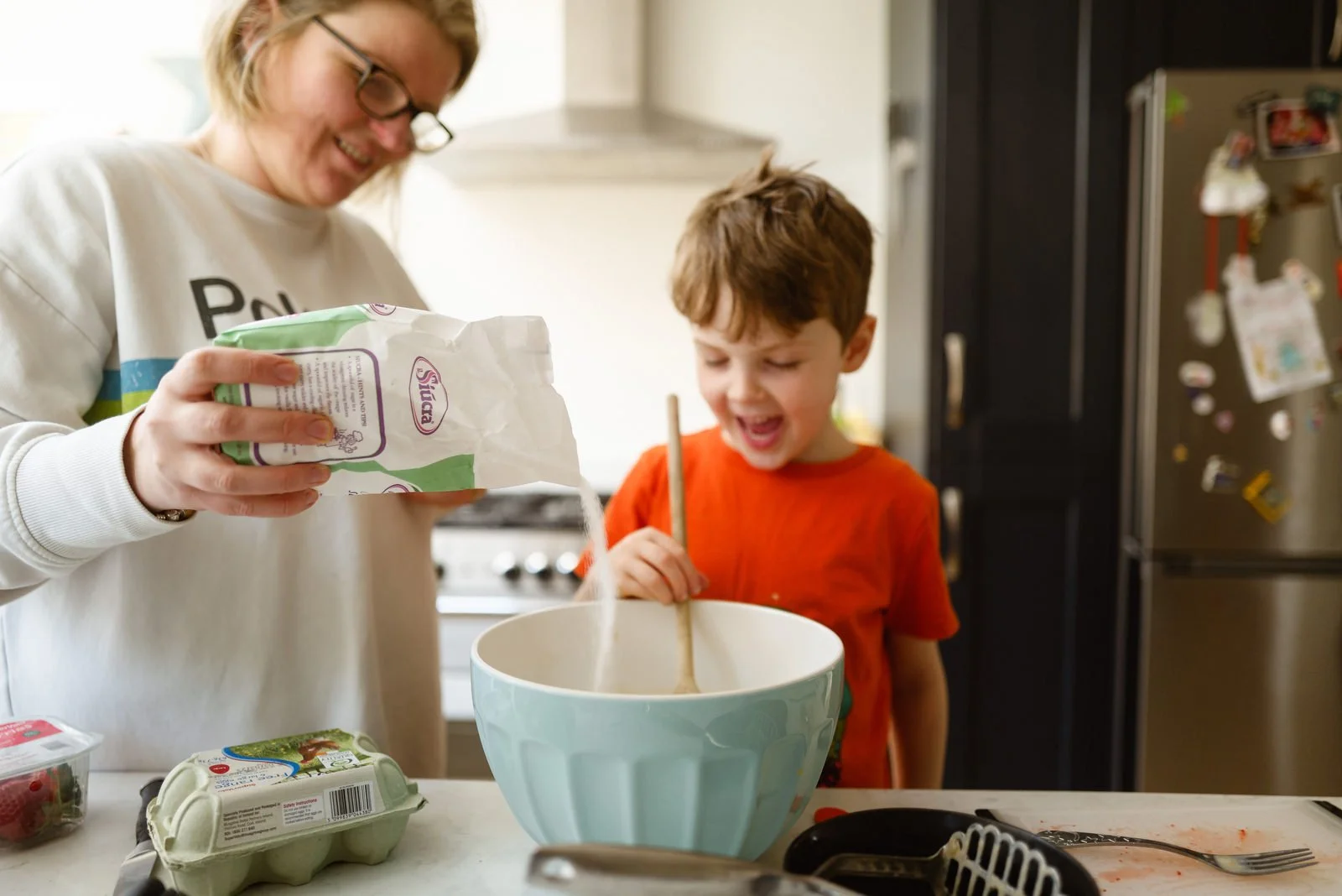 A woman and a young boy in a kitchen baking together, with the woman pouring flour into a mixing bowl and the boy stirring inside the bowl, both smiling.