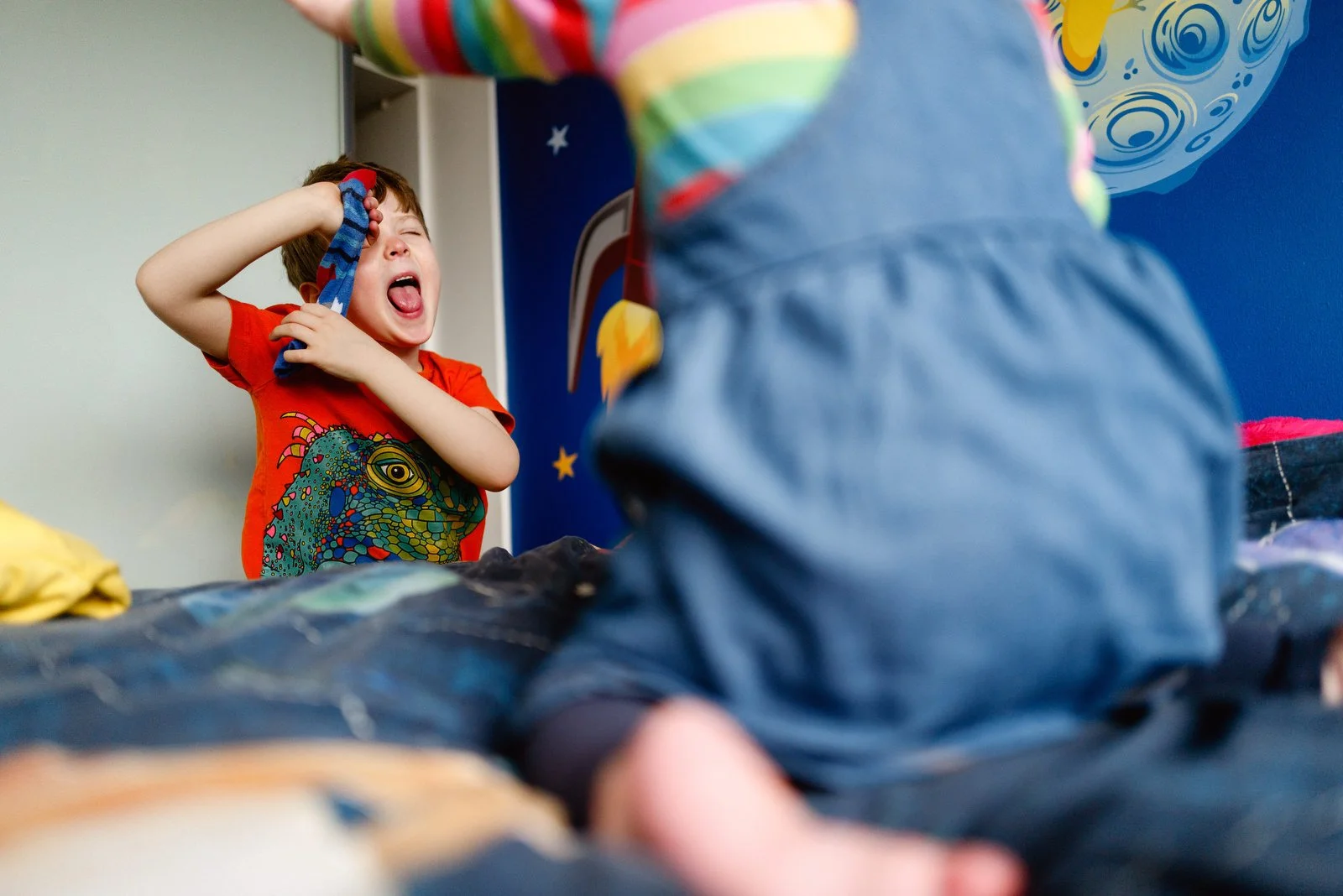 A young boy in a red t-shirt with a monster graphic is playing a game with a person in colorful striped clothing. The boy is laughing and holding a game controller with a playful expression, while the other person is partially visible from the back.