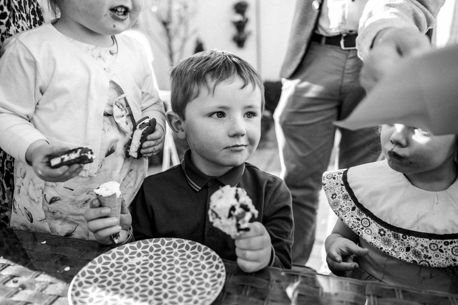A group of young children celebrating a birthday, with one boy sitting at a table holding an ice cream cone, surrounded by girls also eating ice cream. They are in a room with party decorations.