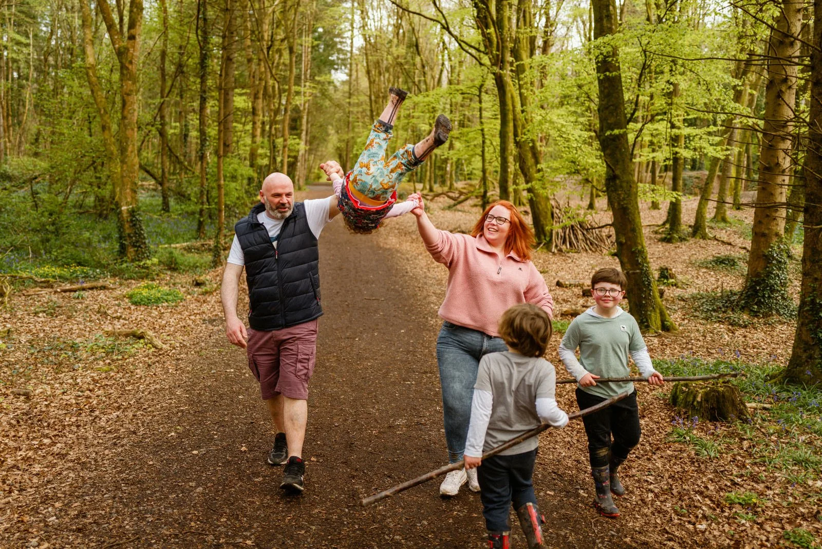 Family of four in a forest, with middle-aged man and woman holding a child's hands as they lift the child into the air, while two kids with sticks walk nearby.