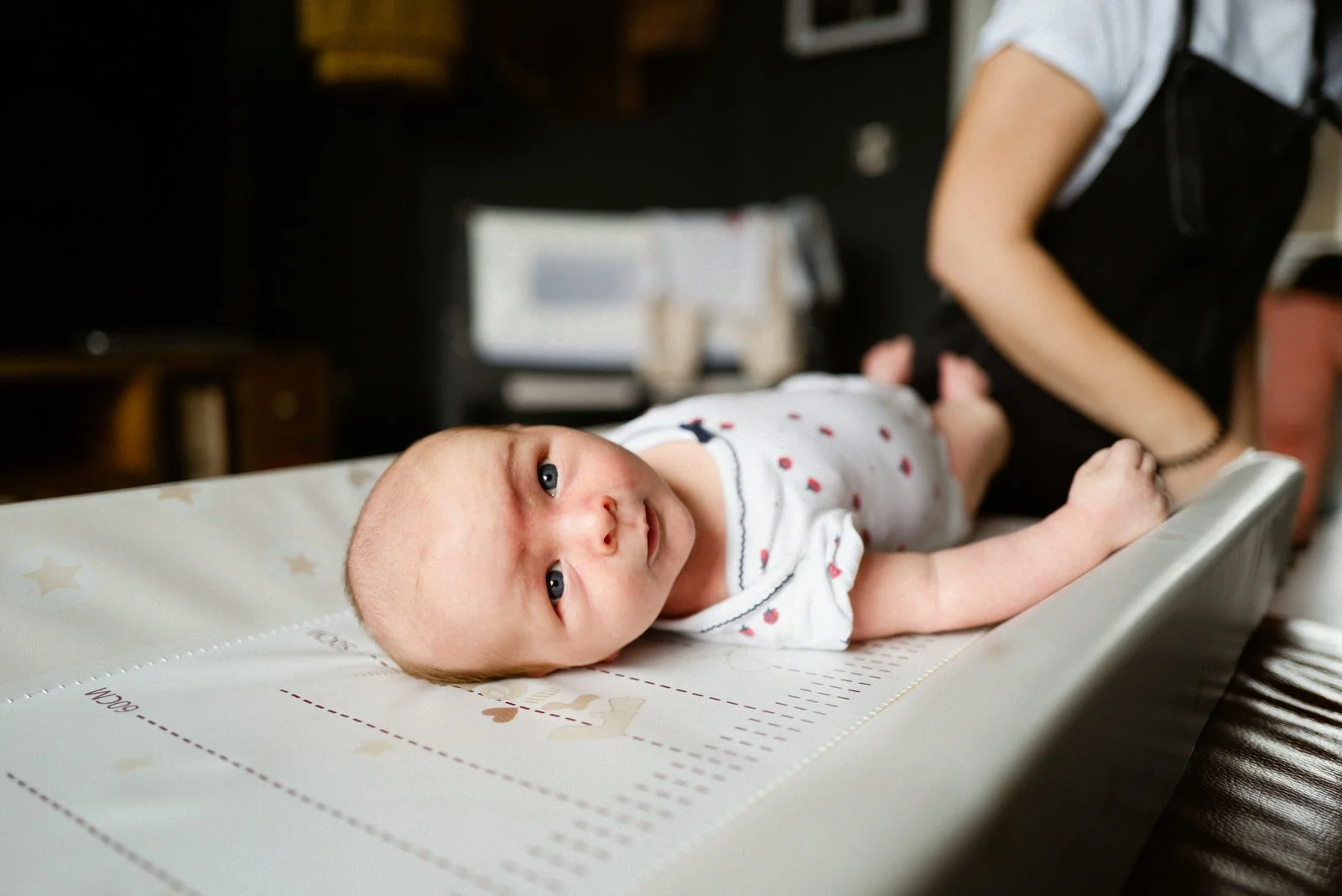 A baby lying on a changing pad with stars, wearing a white onesie with red hearts, looking at the camera.