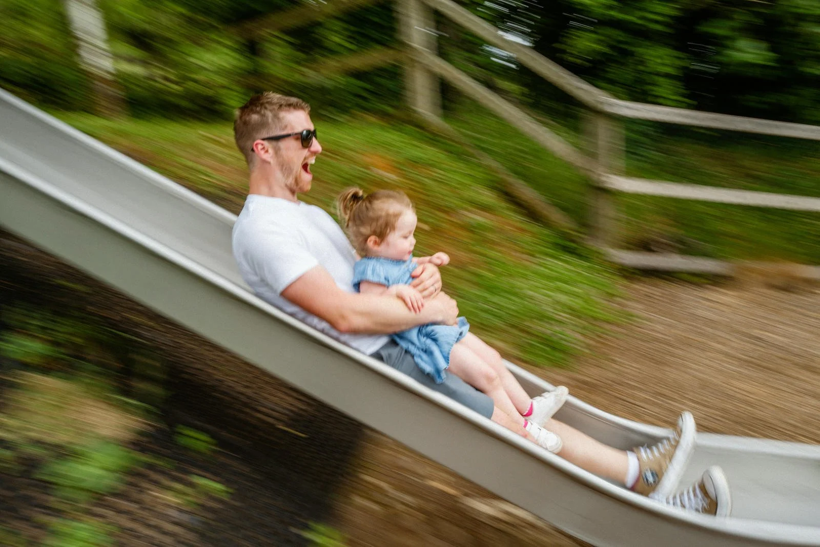 A man wearing sunglasses is riding a slide with a young girl, both enjoying the amusement park ride as the background appears blurred with green trees.