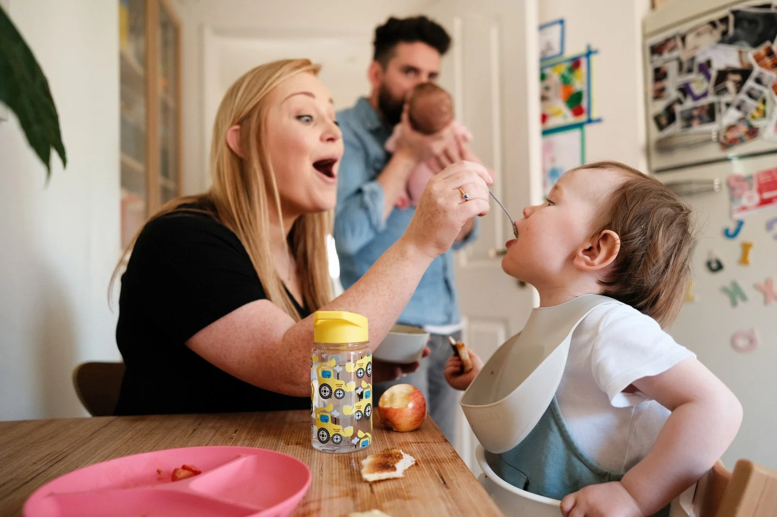 A young girl being fed by a red-haired woman at a kitchen table, with a man in the background holding a baby. Expressions indicate surprise or amusement.