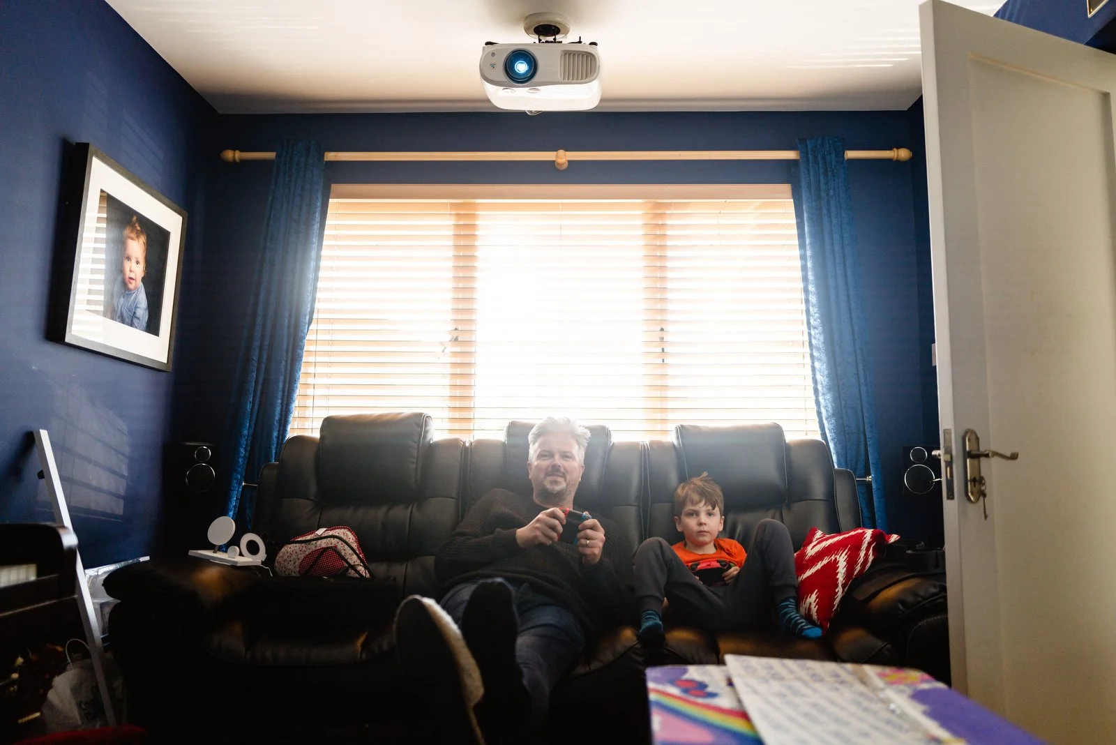 A man and a young boy sitting on a black leather couch in a living room, with bright sunlight coming through large windows with blue curtains behind them. A white ceiling-mounted projector is visible at the top of the image.