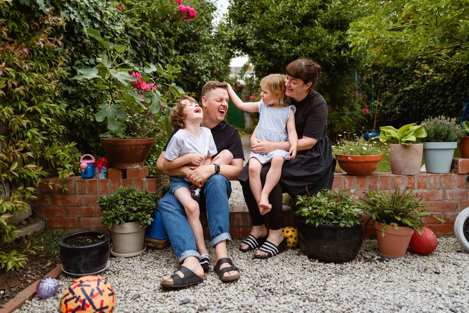 A family of four sitting outdoors on a brick wall surrounded by potted plants and garden decorations, laughing and enjoying each other's company.