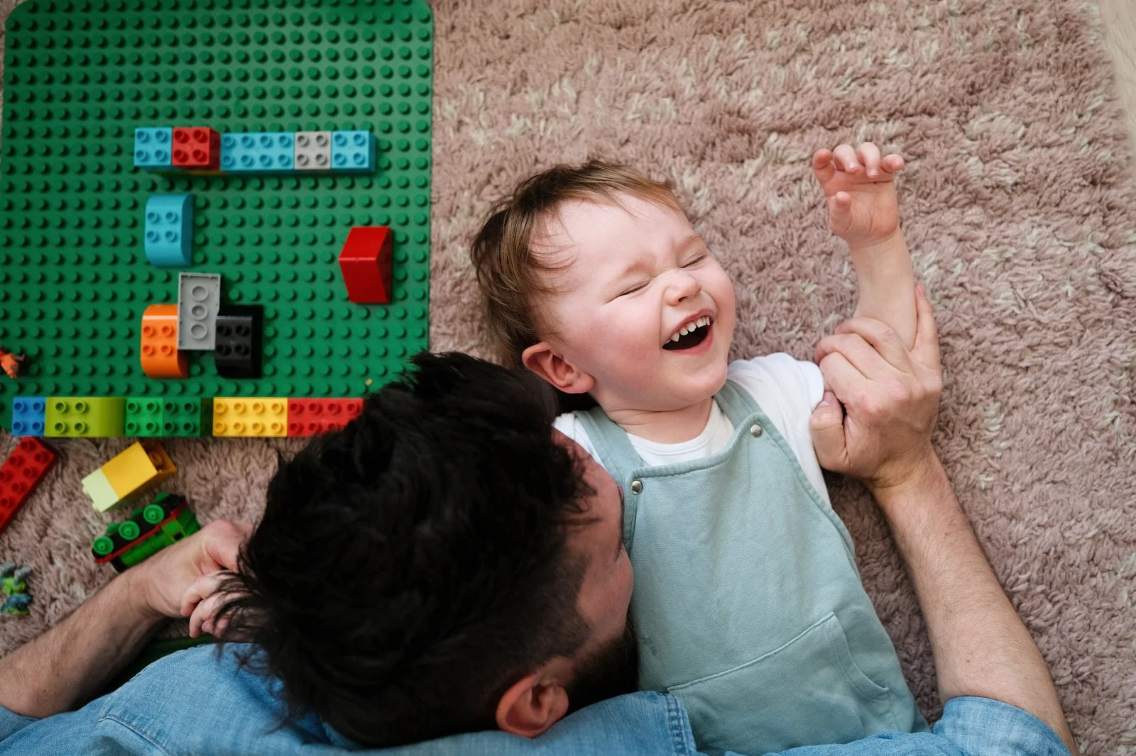 A young boy with Down syndrome laughing and lying on a carpeted floor, hugging an adult with dark hair, next to a green Lego baseplate and scattered colorful Lego bricks.