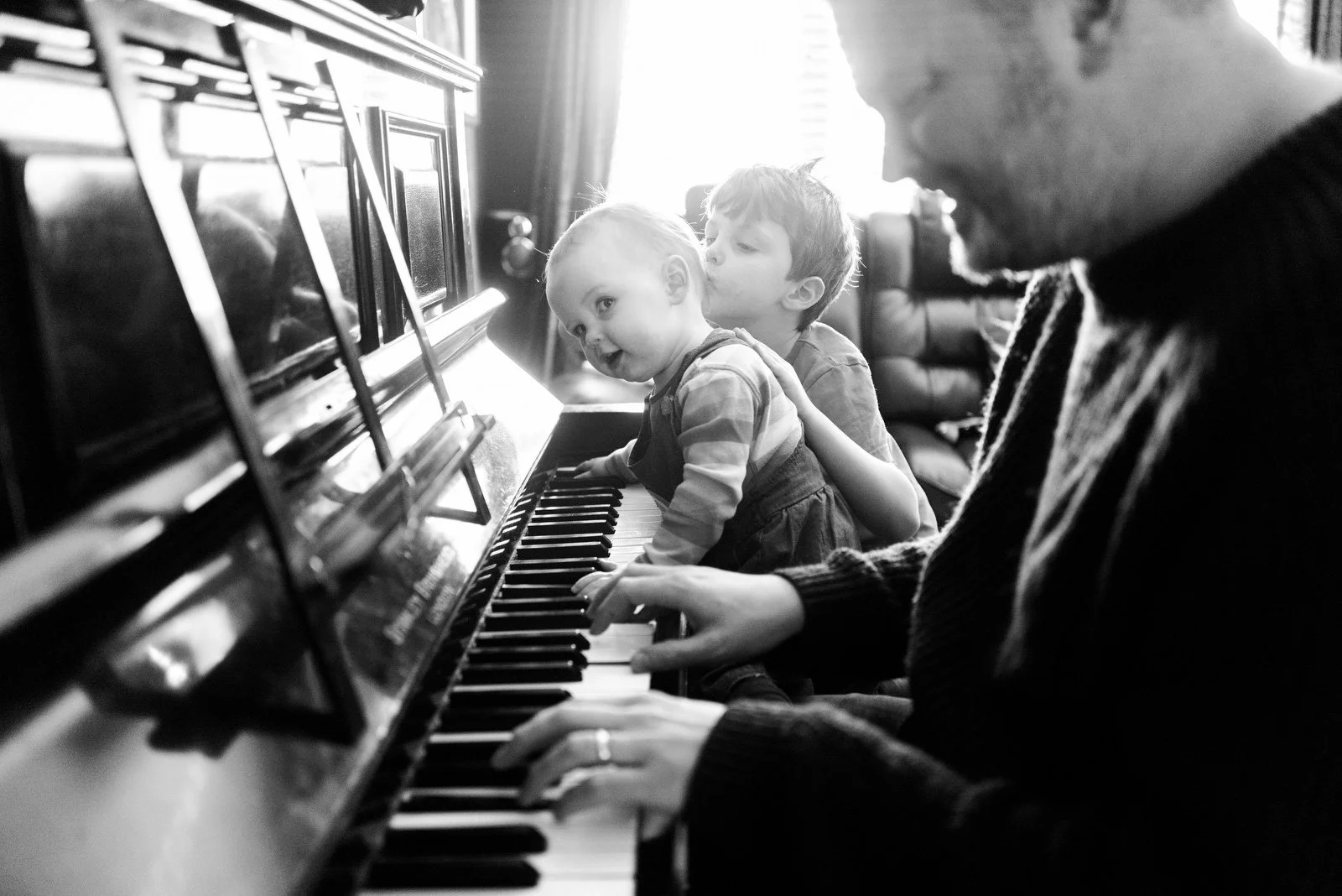 A person playing the piano with two children sitting on the bench, one of them touching the keys, and the younger child looking at the camera, in a room with natural light.