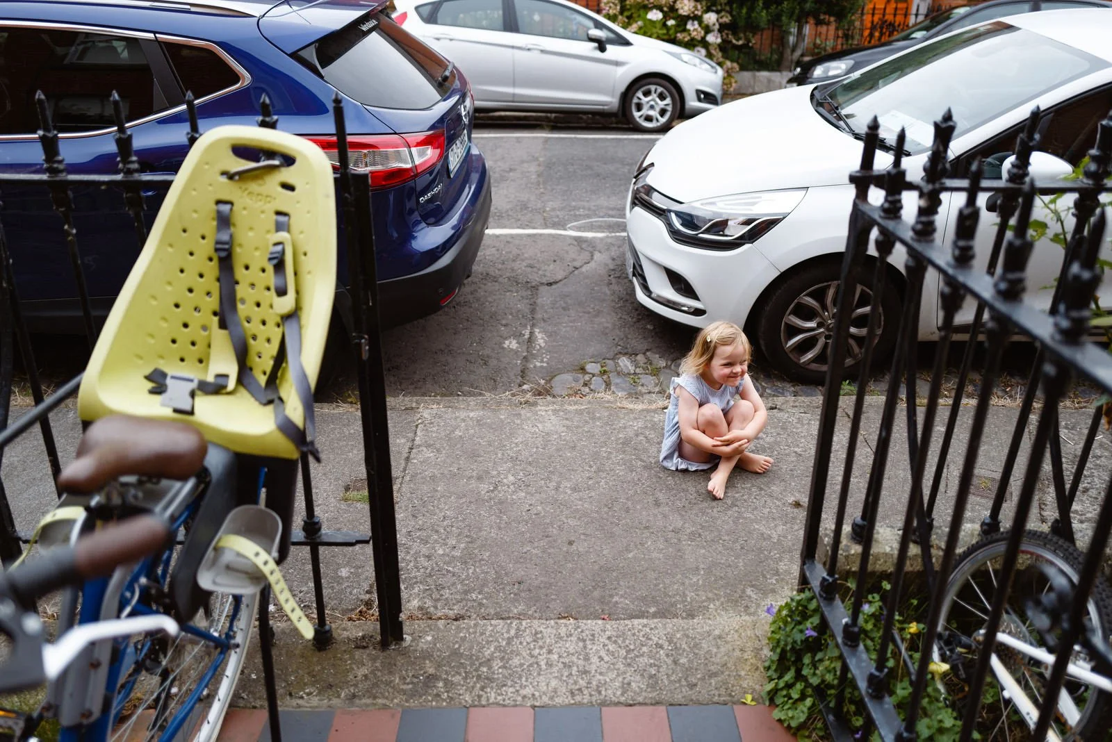 A young girl sitting on a sidewalk outside, smiling and hugging her knees, with cars parked along the street behind her and bicycle storage with a child's seat in front of her.