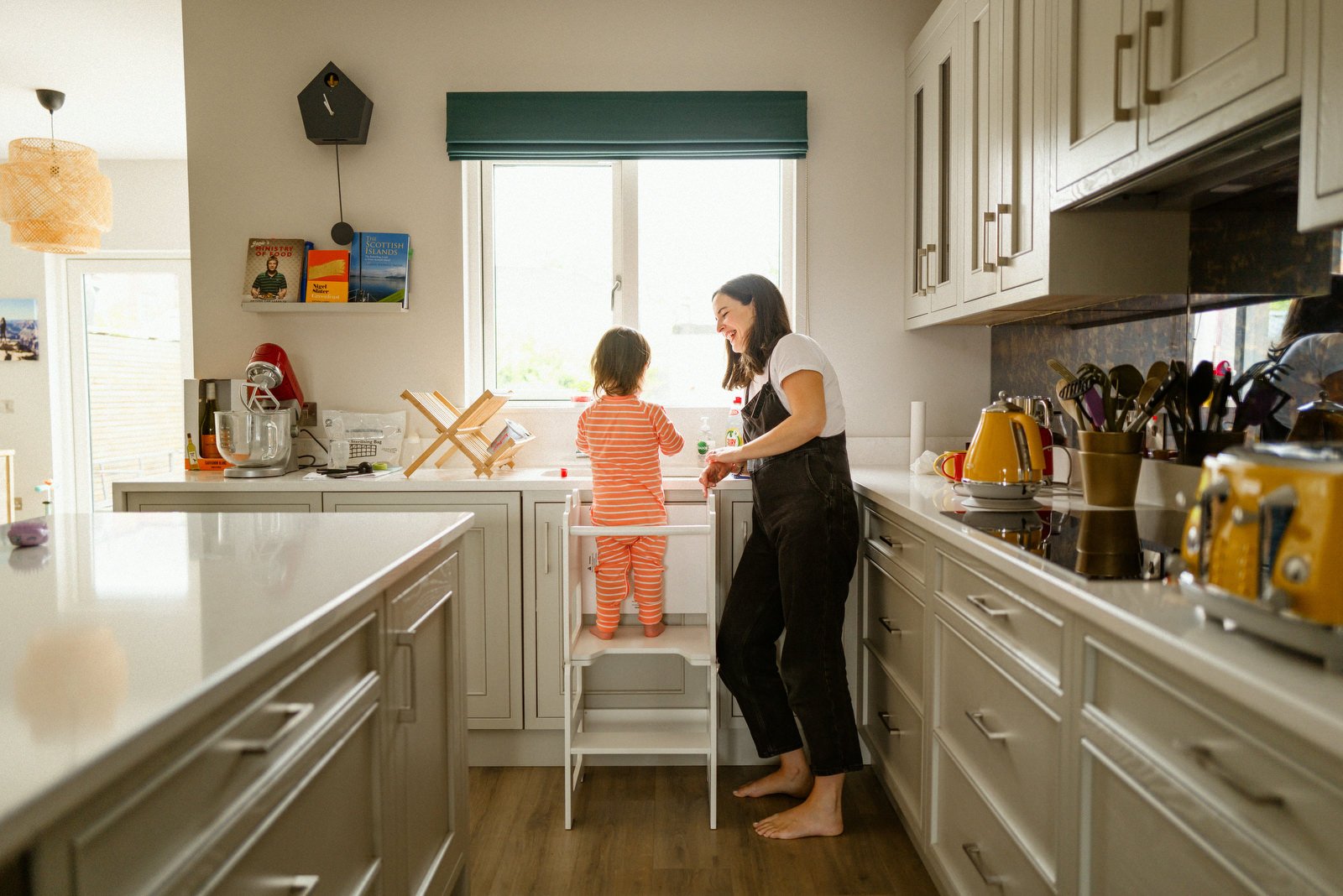 A woman and a young girl are in a kitchen; the girl is standing on a step stool by the sink, and the woman is smiling and interacting with her, near a window, with various kitchen appliances and items around.