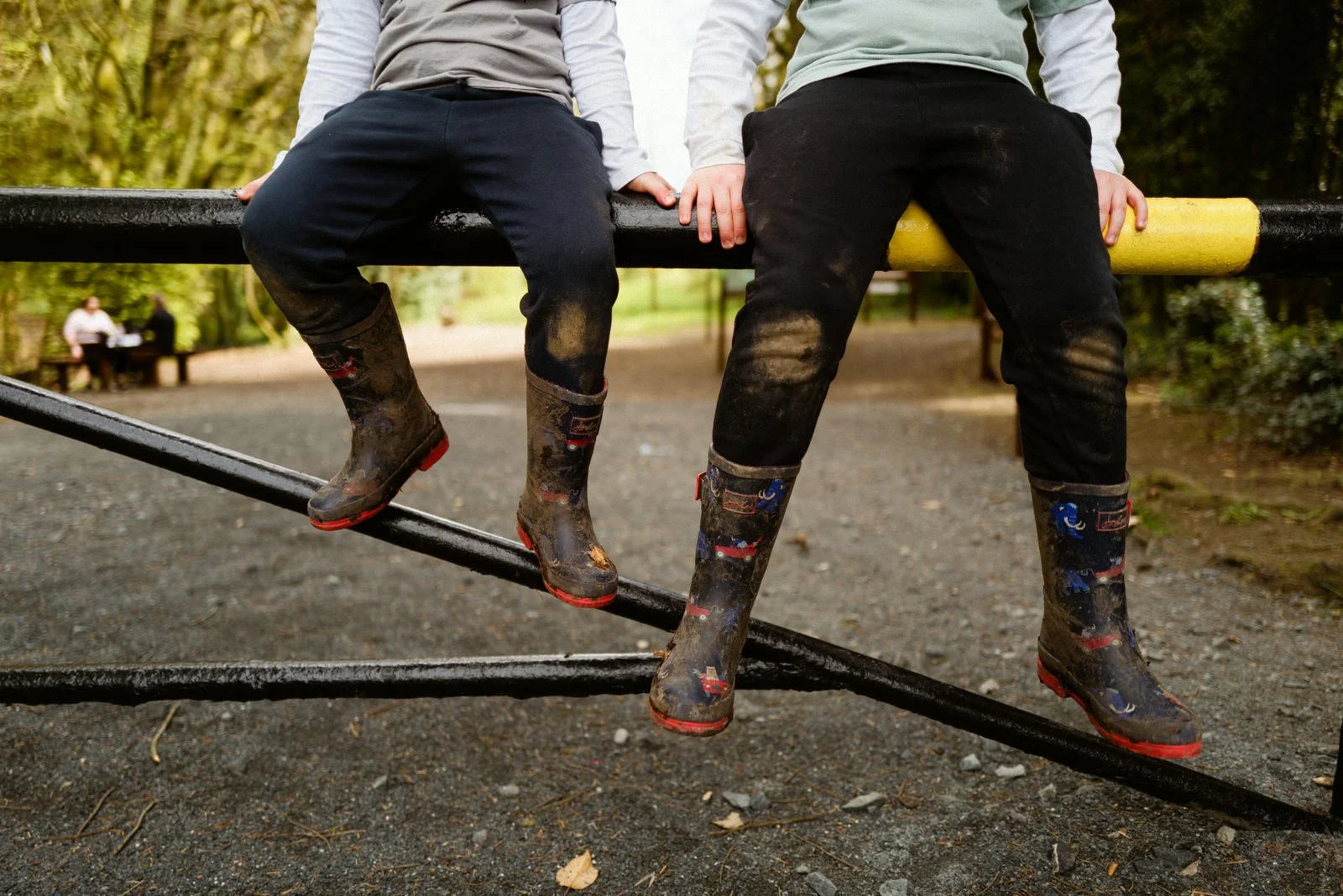 Two children sitting on a park fence, wearing muddy boots and pants, with a park scene in the background.
