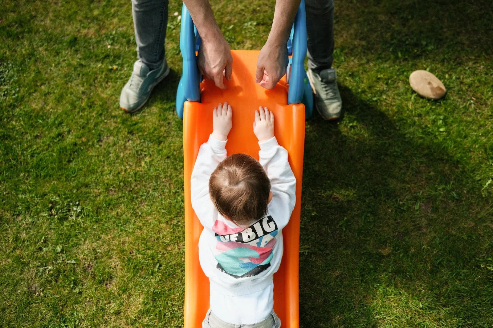 A child lying face down on an orange slide, being held by two adults at the top of the slide outdoors on grass.