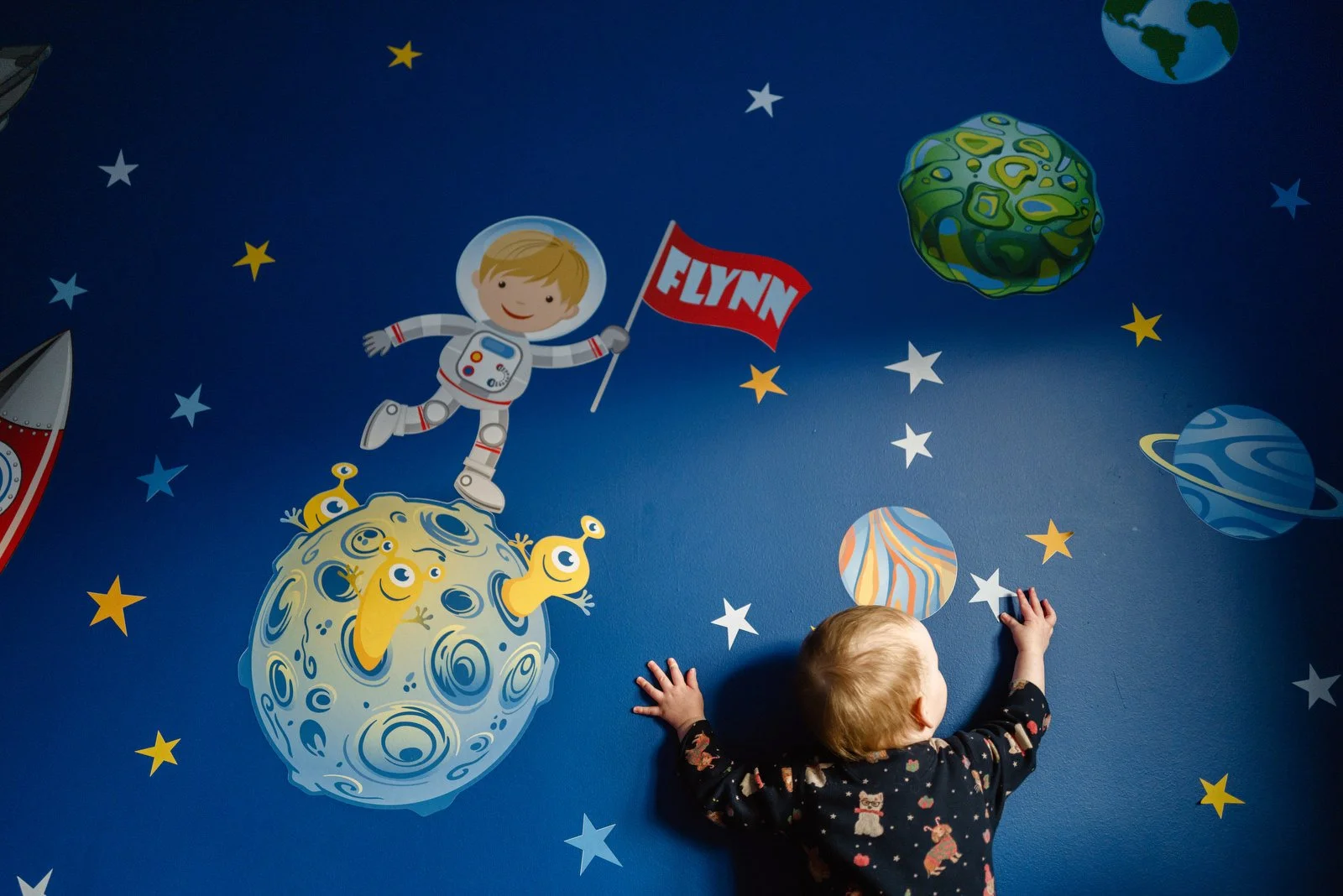 Child in pajamas playing in front of a space-themed wall mural with cartoon planets, stars, and an astronaut figure holding a 'FLYNN' flag.