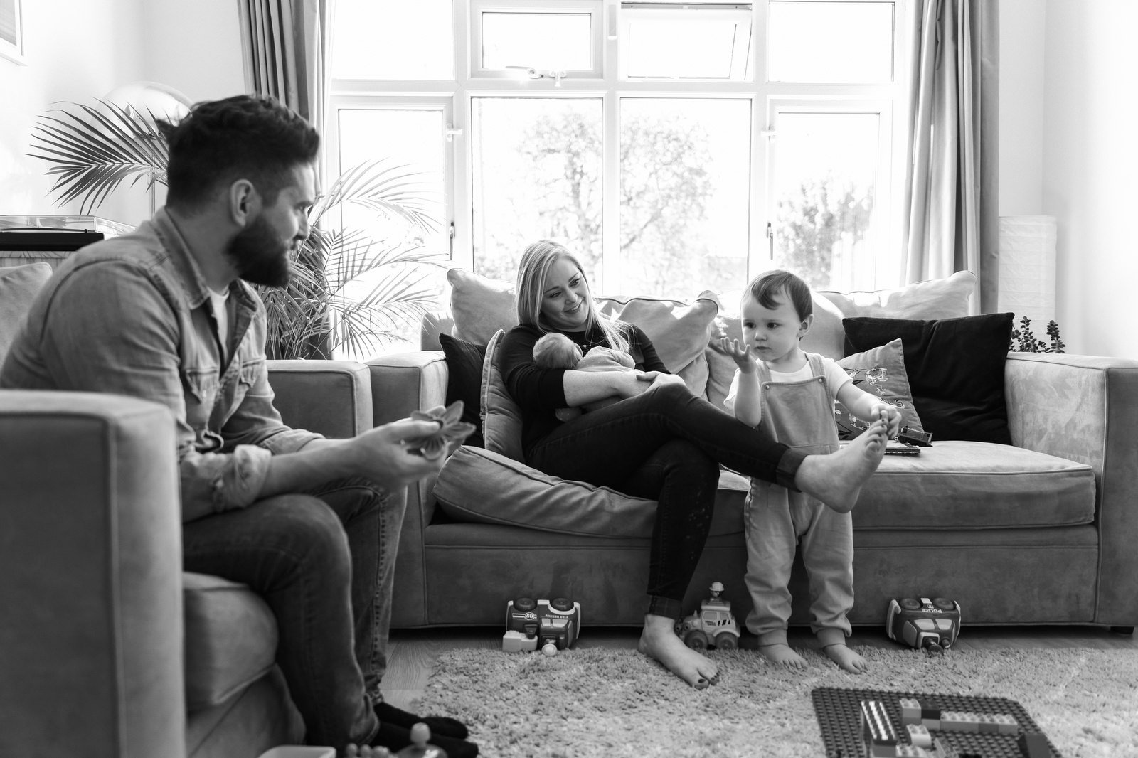 A family of four sitting on a sofa in their living room, with a man on the left holding a toy and a woman in the middle holding a baby. A young girl standing barefoot next to the sofa is gesturing with her hands. Toys are on the floor and the room is