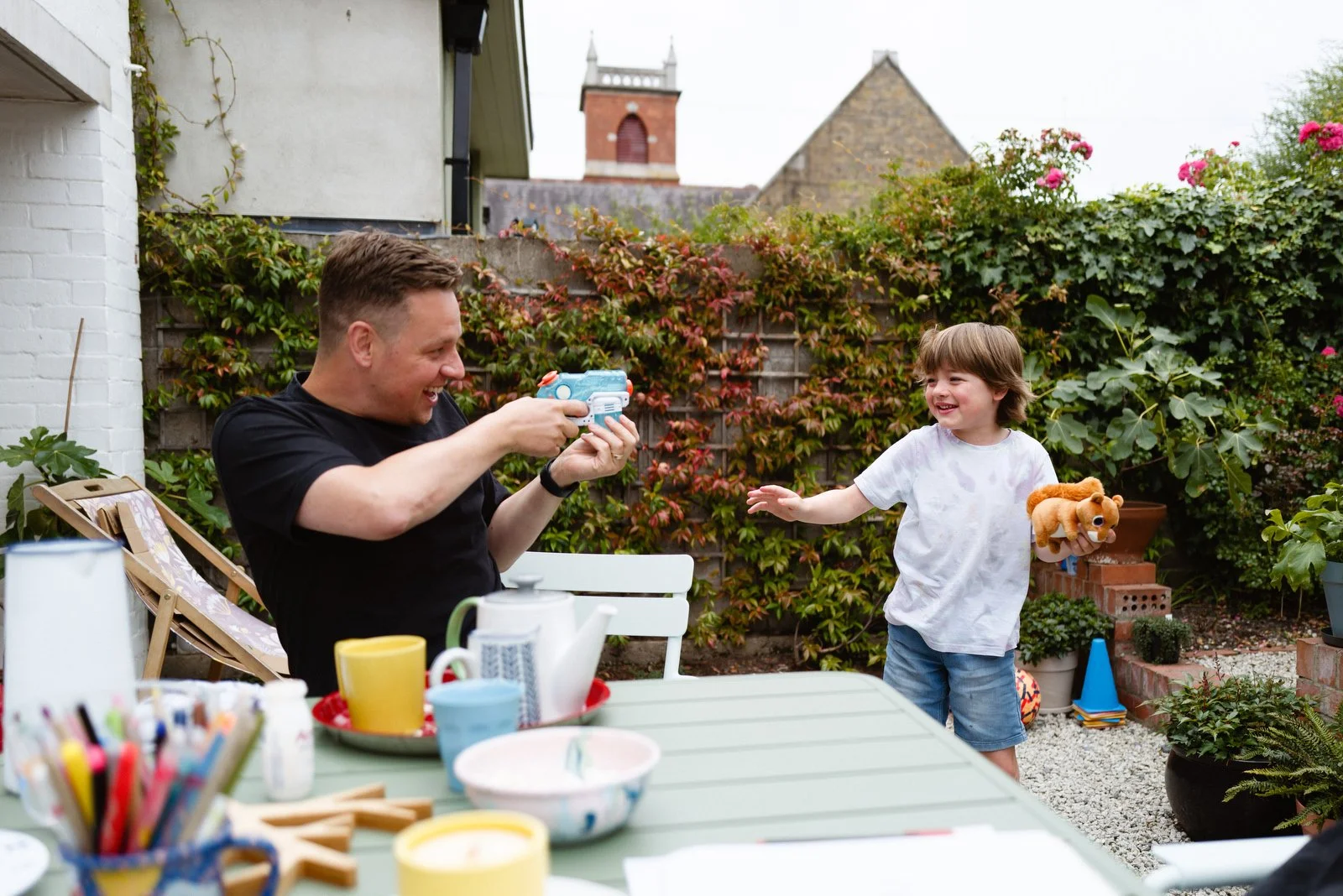 A man and a young boy in a garden, playing with toy guns and laughing. The boy is holding a plush toy.