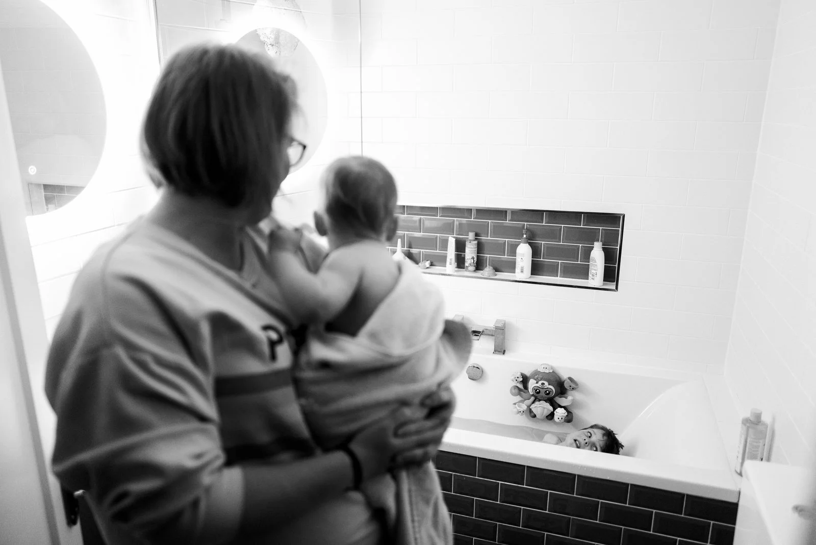 A woman holding a child near a bathtub with a child inside with toys, in a bathroom with shelves and toiletries.