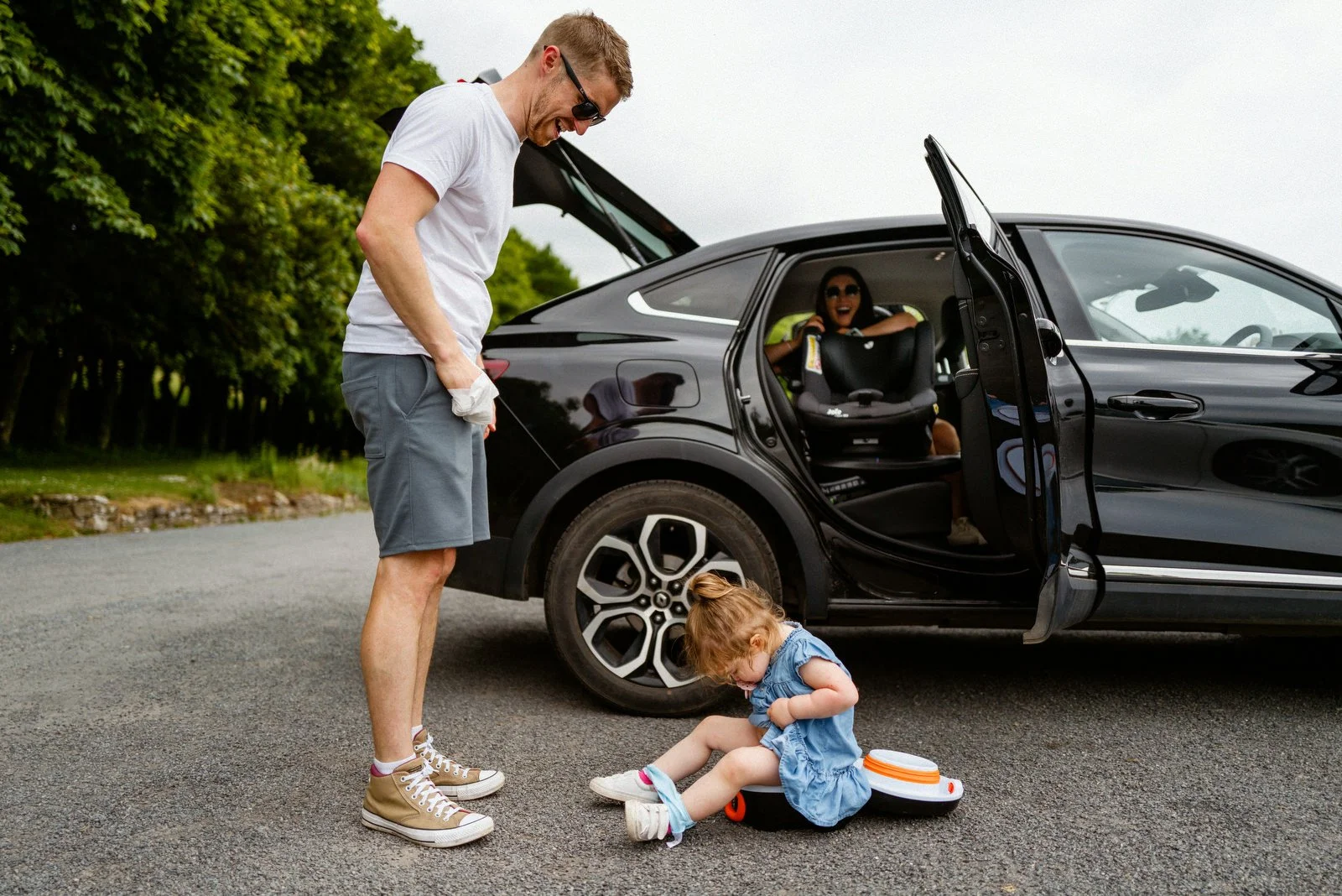 A man in casual clothing standing outside a black car while a young girl is sitting on the ground fixing her shoe, with a woman laughing in the backseat of the open car door.