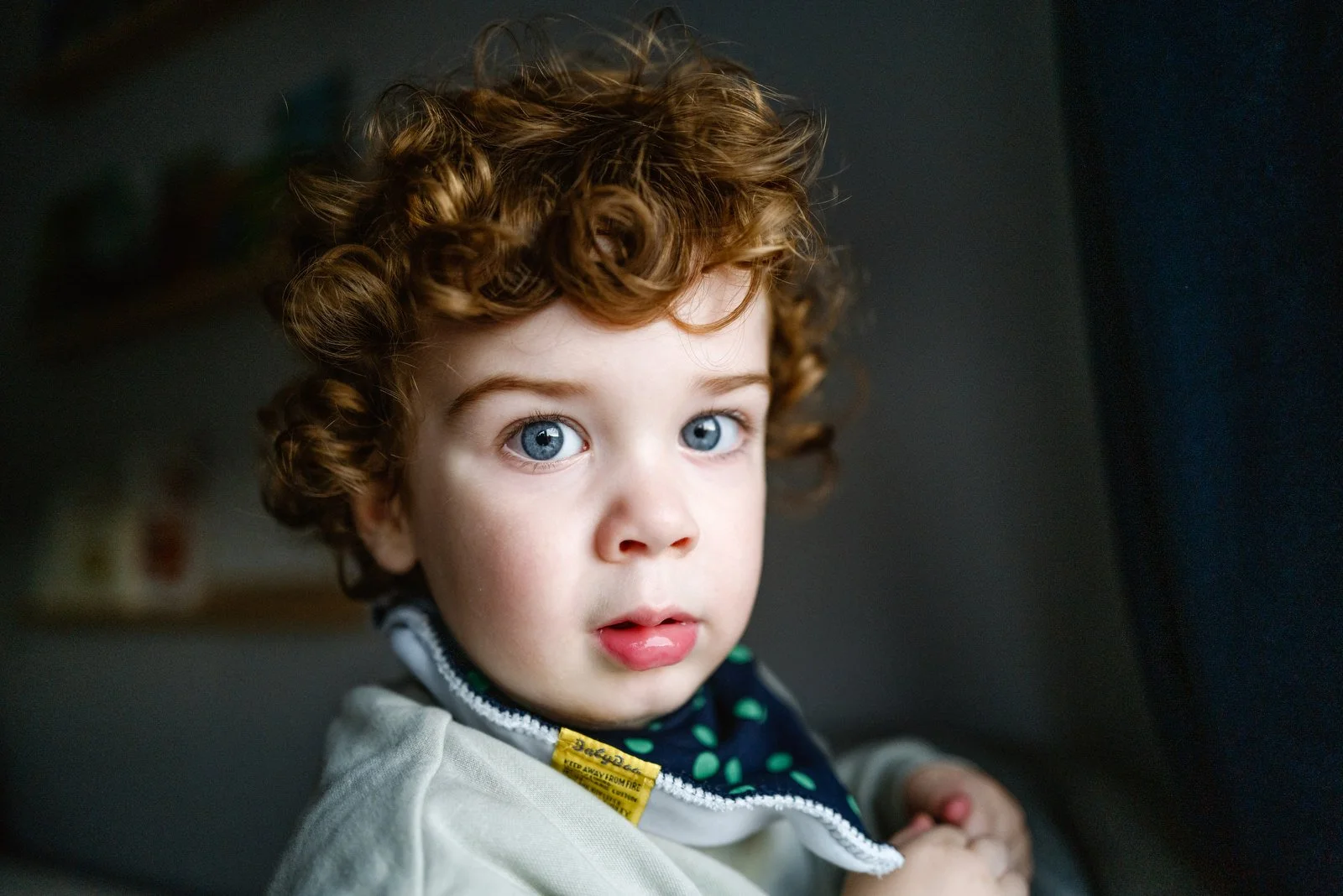 Close-up of a young child with curly red hair and blue eyes, wearing a light-colored jacket and sitting indoors.