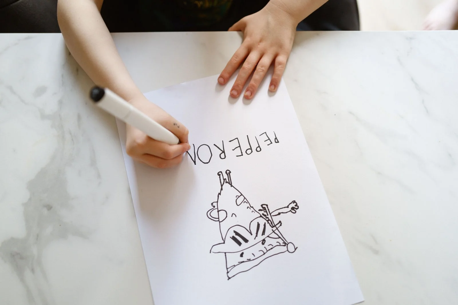 A child is drawing a cartoon slice of pizza with a face on a white sheet of paper while writing the word "PIZZA" in large letters. The child is holding a black marker and has one hand resting on the paper.