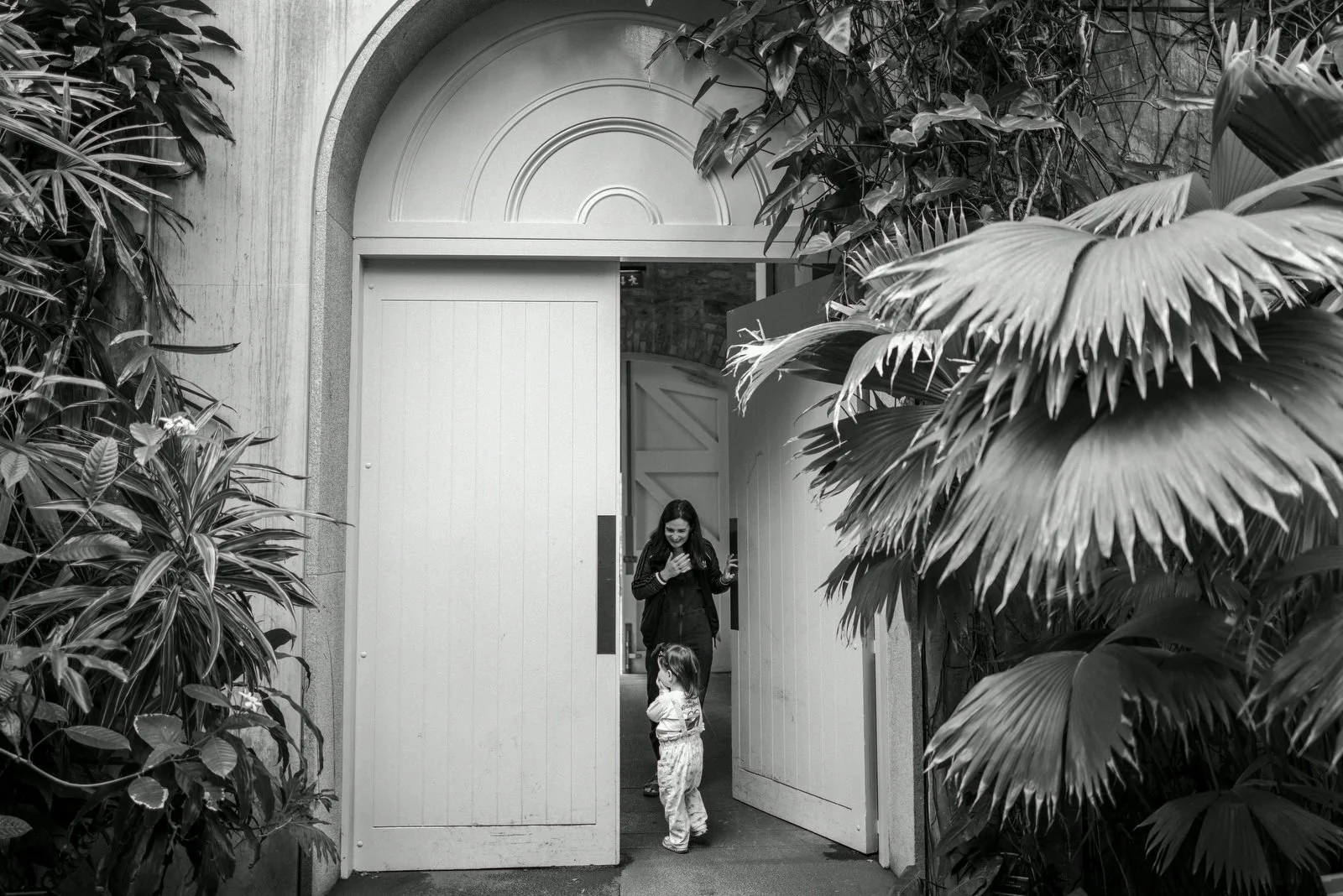 A woman and a young girl standing in a doorway surrounded by lush tropical plants. The woman is smiling and looking down at the girl, who is facing her.