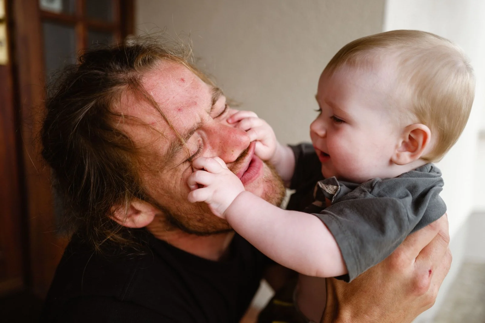 A man with long hair and a beard holding a young child with blonde hair, both smiling and playing with each other's faces.