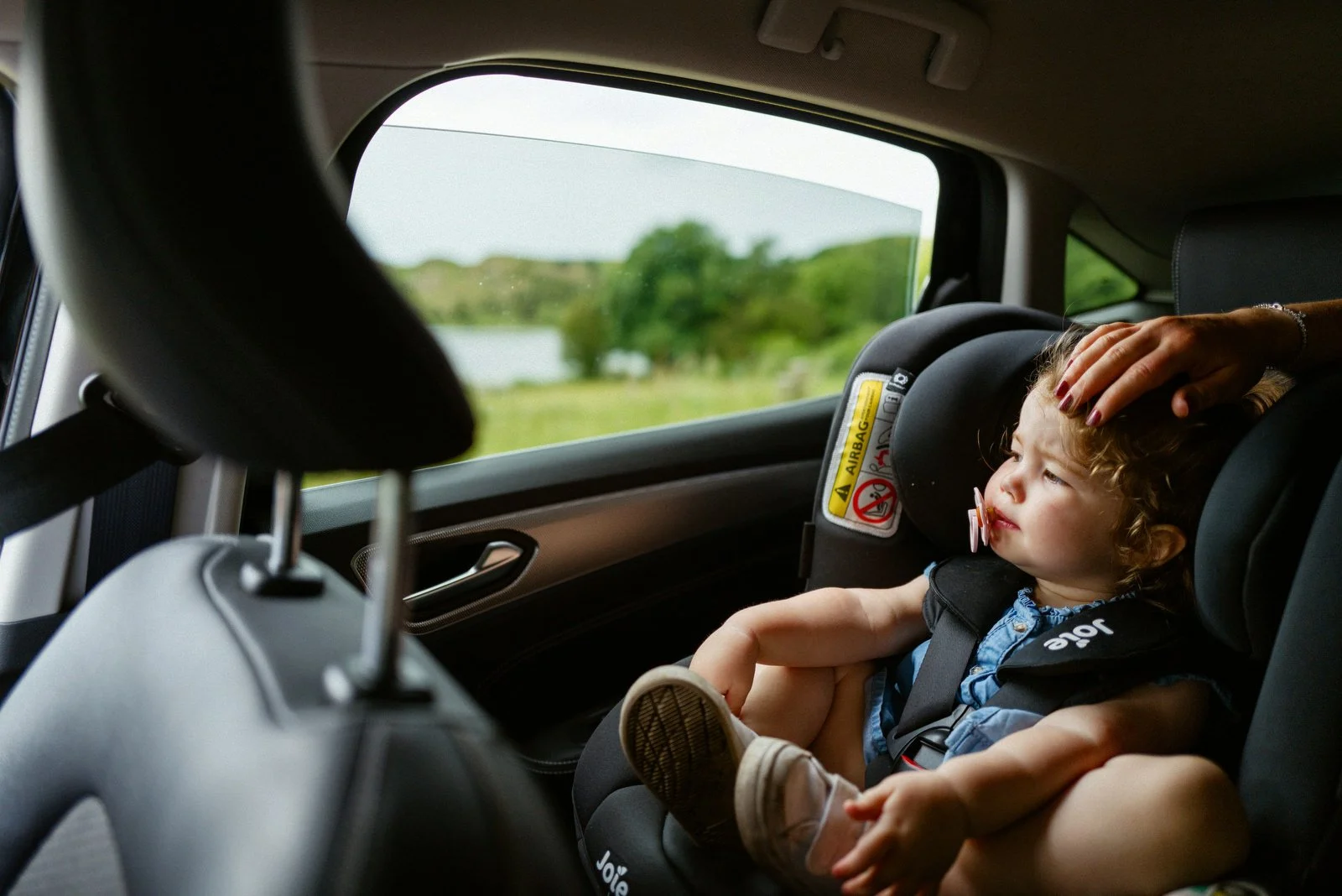 A young child with curly hair sitting in a car seat inside a vehicle, with an adult's hand resting on the child's head, looking out the window at a scenic view of trees and water.