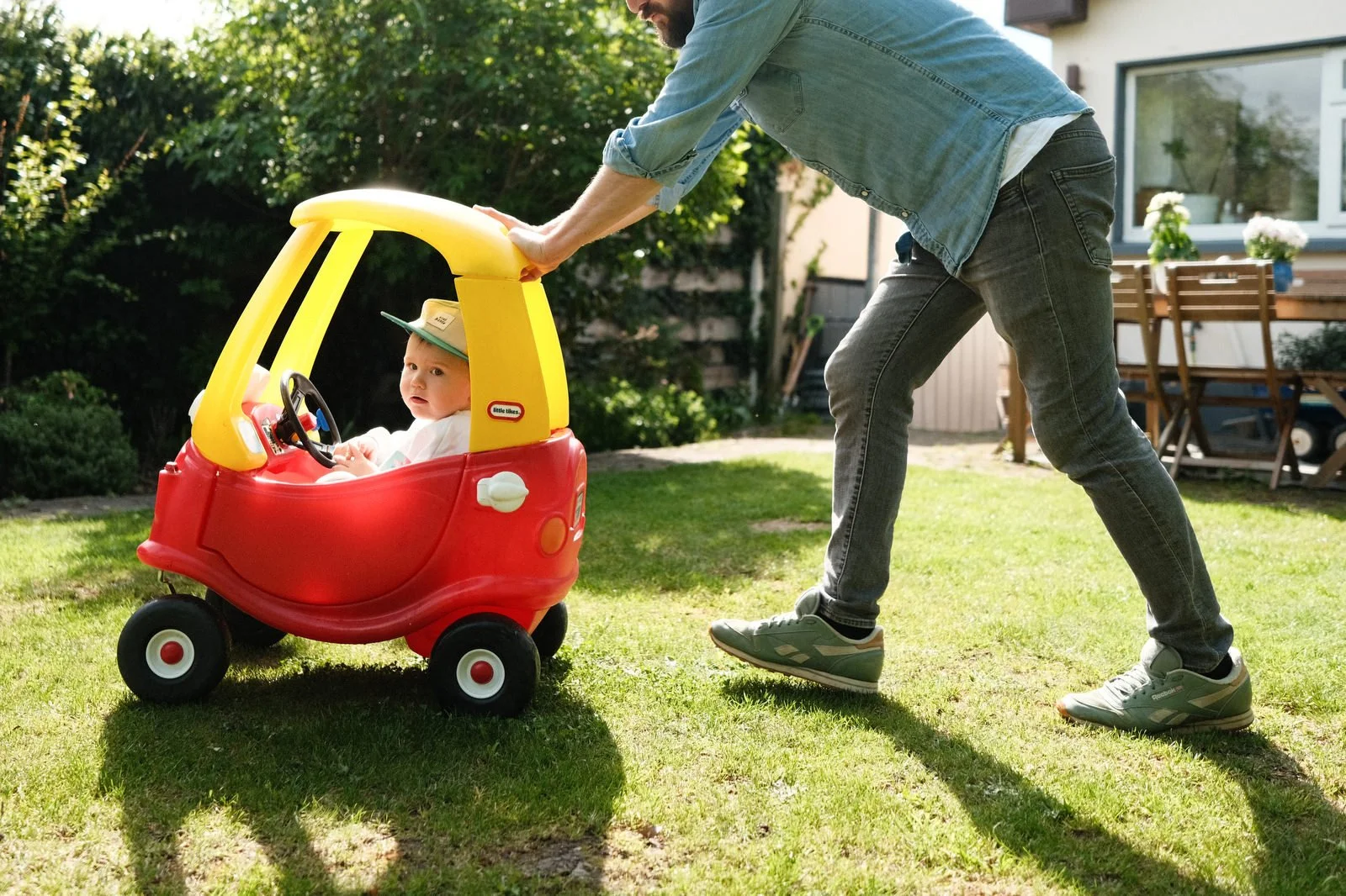 A man pulling a toddler in a red and yellow toy car in a backyard on a sunny day.