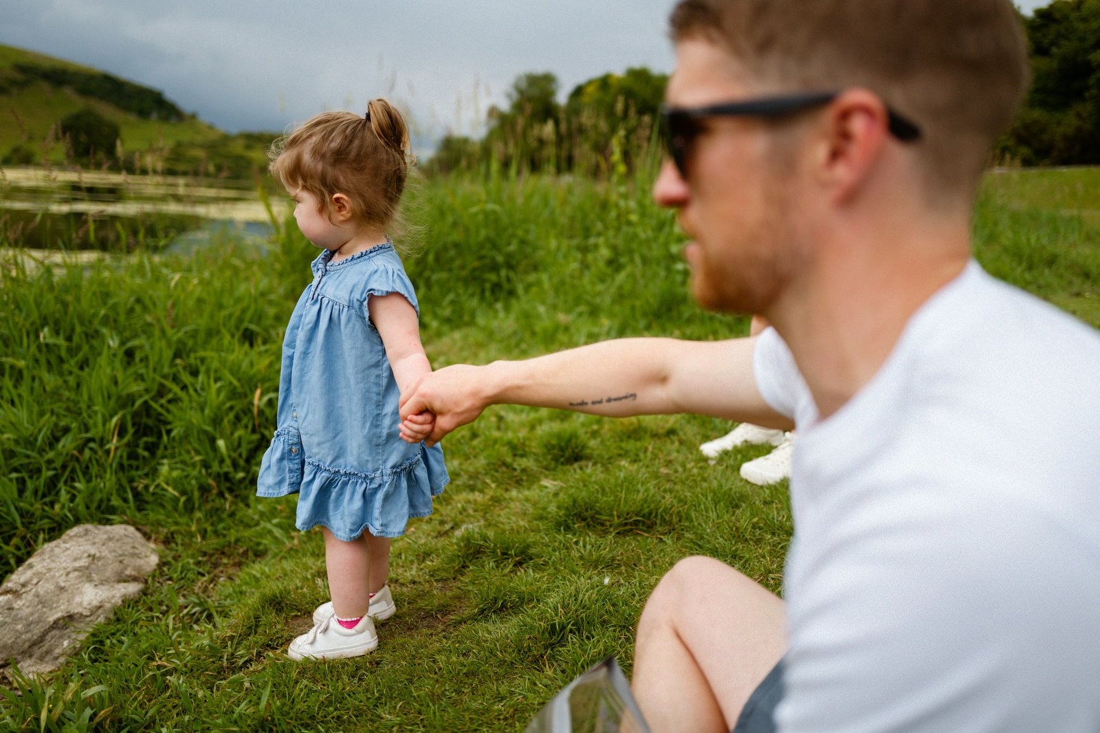 A young girl in a blue dress holding hands with a man sitting outdoors near a lake, with green hills in the background.