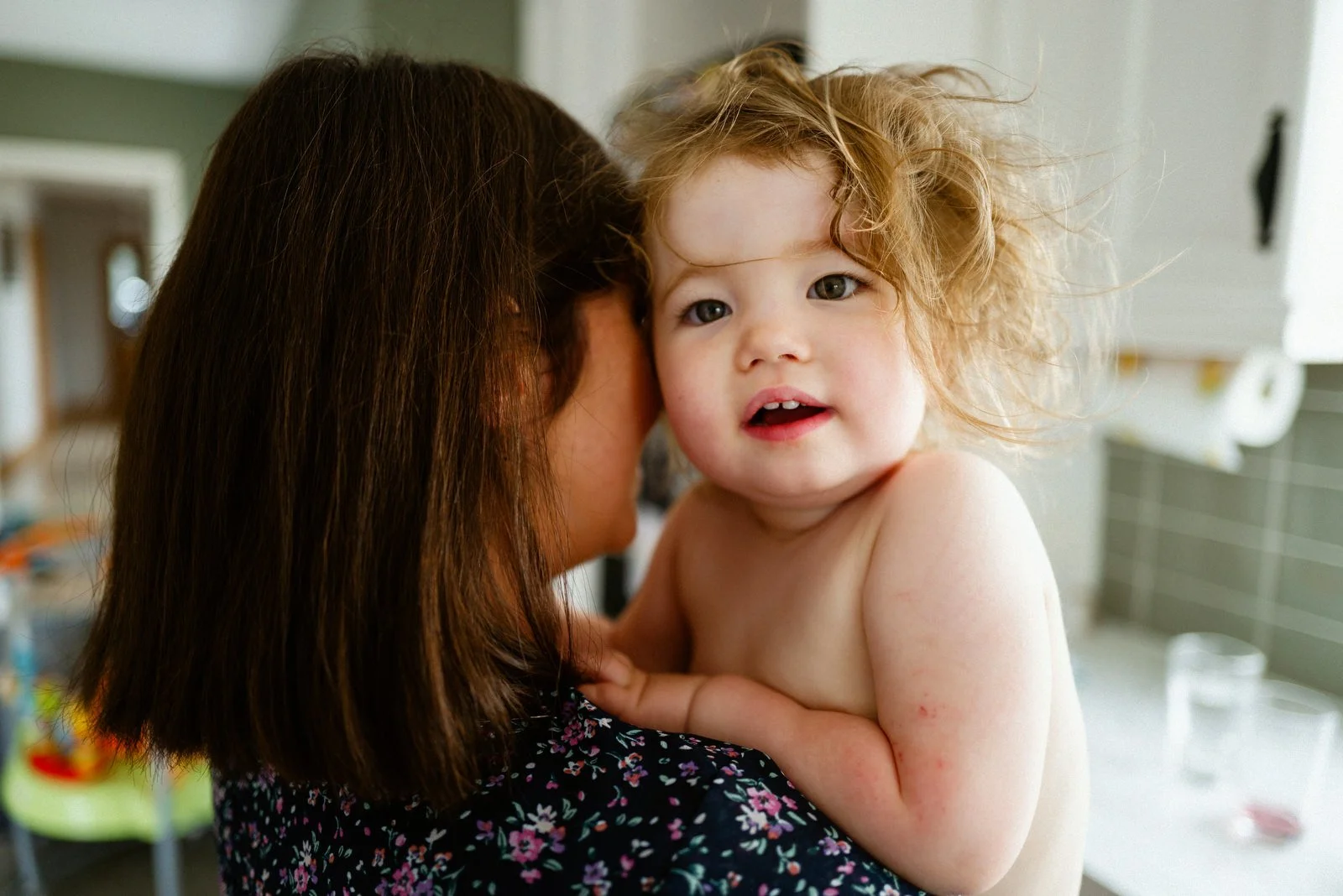 A young child with curly red hair looking at the camera, being held by an adult with brown hair in a domestic setting.