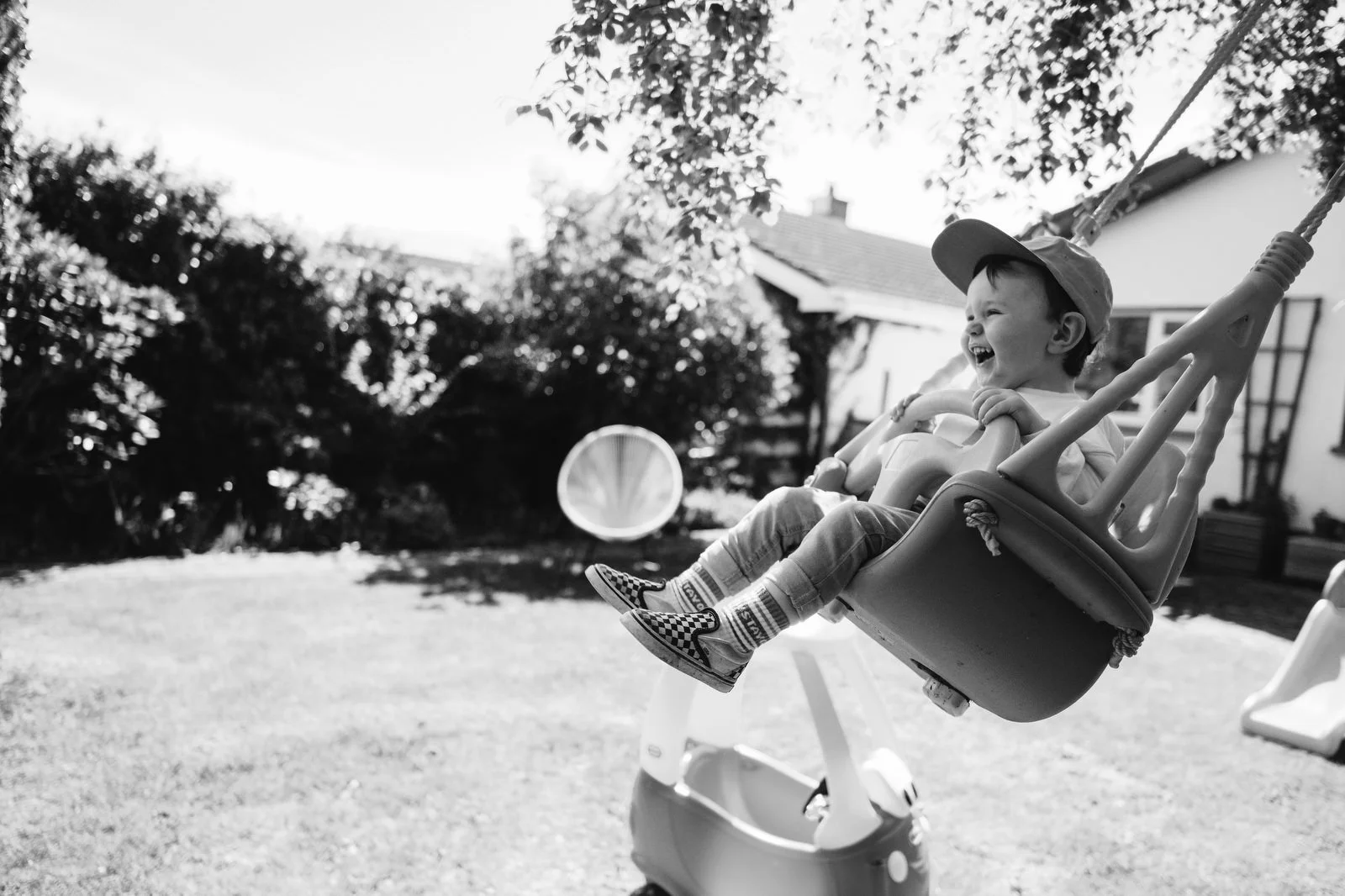 A young boy wearing a cap, sitting in a swing, laughing and smiling outdoors on a sunny day.