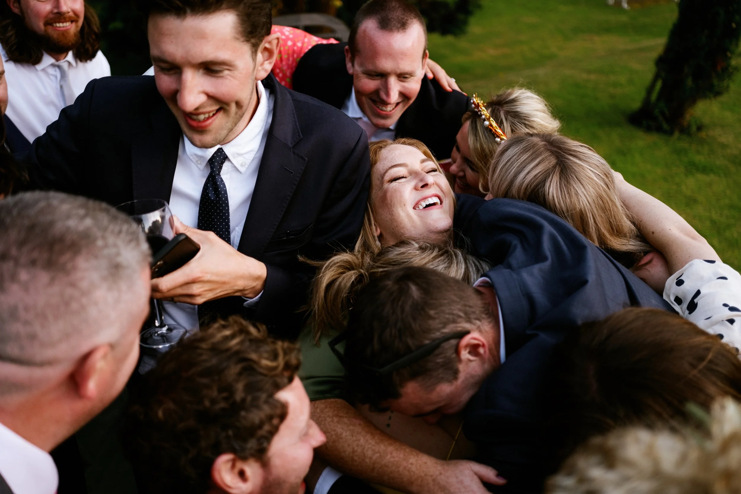 A group of people dressed in formal attire are hugging and laughing outdoors on a grass lawn during a celebration.
