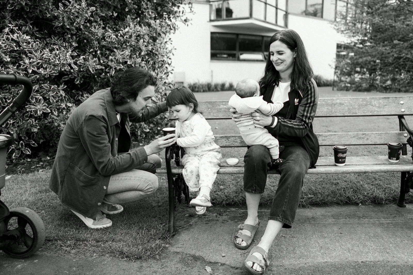 A woman sitting on a bench holding a baby, with a young girl sitting next to her. A man kneeling on the grass is handing a cup to the girl. There are three coffee cups on the bench and a stroller partially visible on the left. The scene is outdoors w