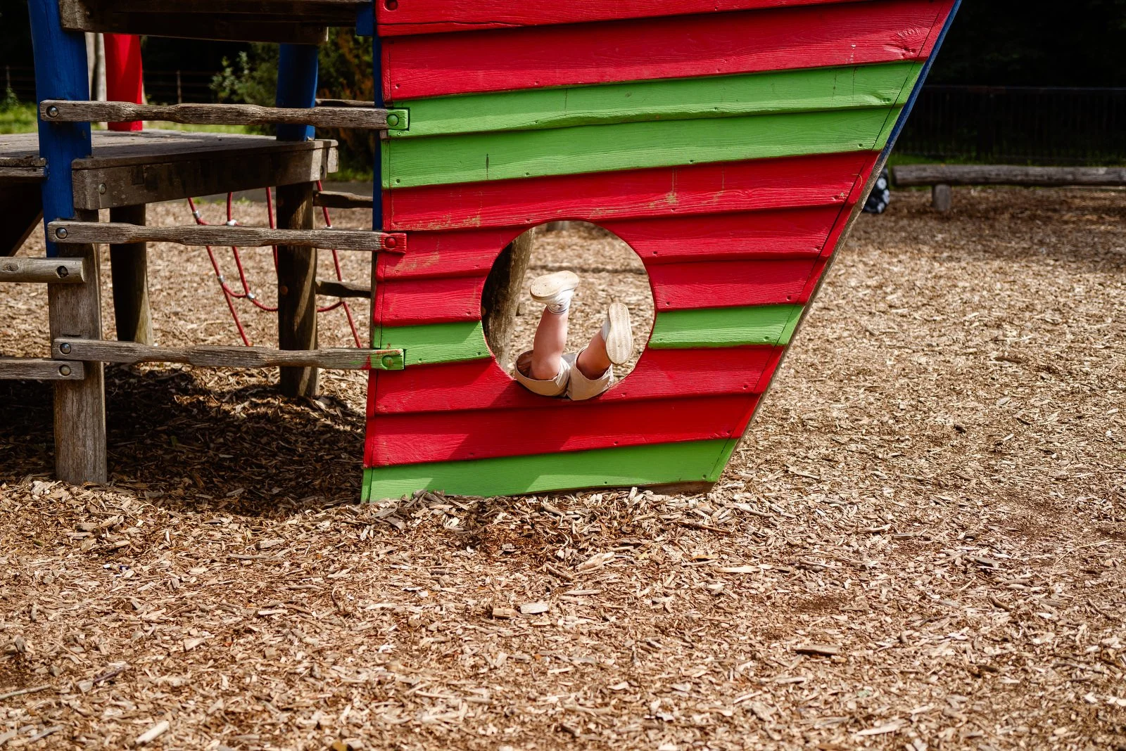 Child crawling through a hole in a colorful wooden playground structure painted red and green, with wood chips on the ground.