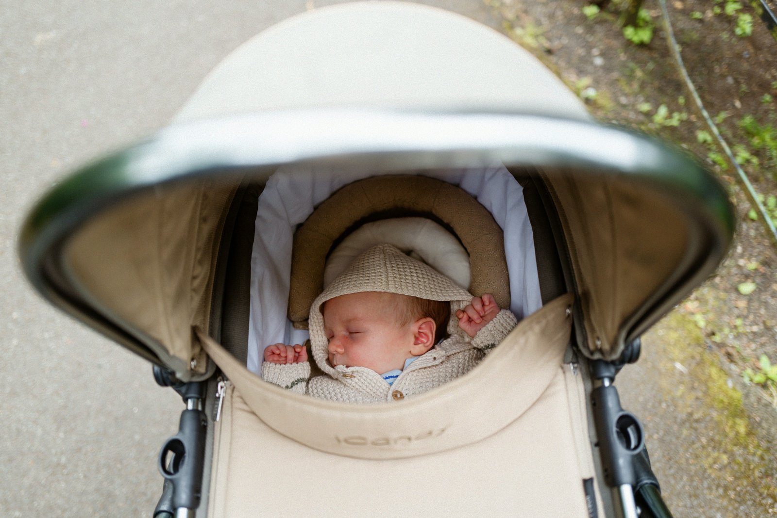 A baby sleeping in a stroller with a beige hood, dressed in a knitted sweater and bonnet, with a circular pillow around its head, outdoors on a concrete and mossy surface.