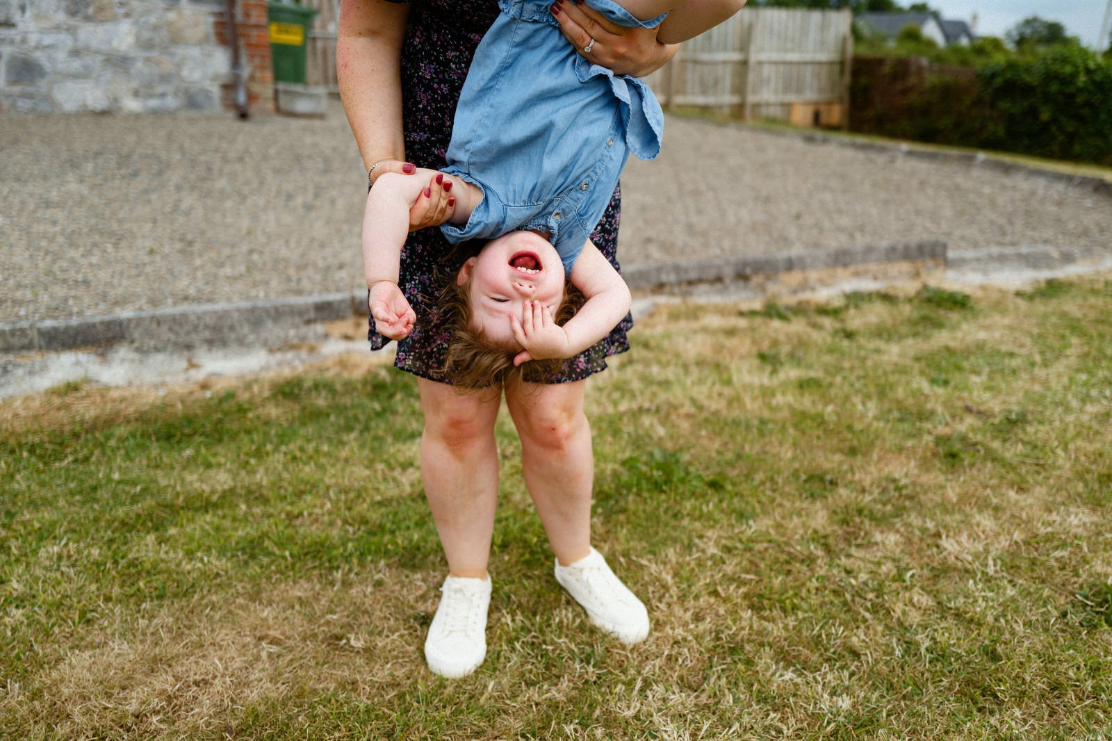 A young girl in a blue dress holding her head, being swung upside down by an adult in a black floral dress in an outdoor yard with grass and a gravel area.