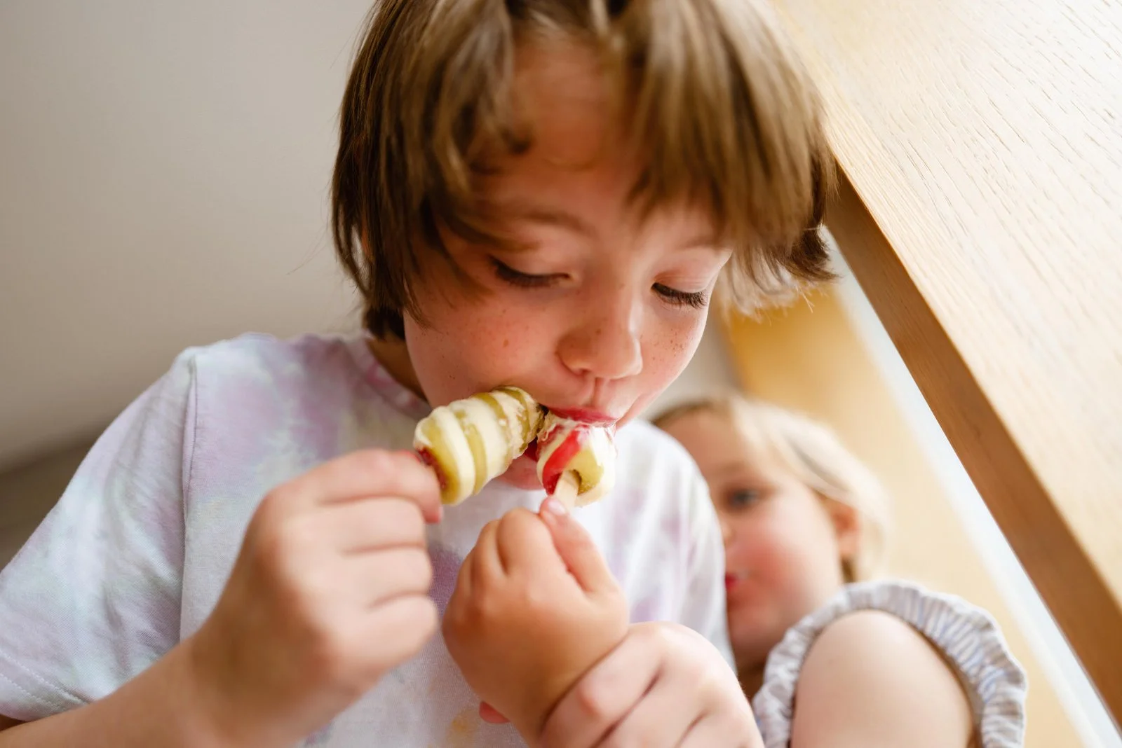 A young boy with brown hair and freckles eating a vanilla and strawberry ice cream cone. A blonde girl in the background is looking on.