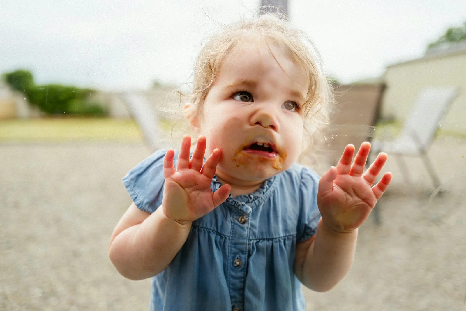 Child looking through a glass door with hands up, face stained with food, wearing a blue dress.