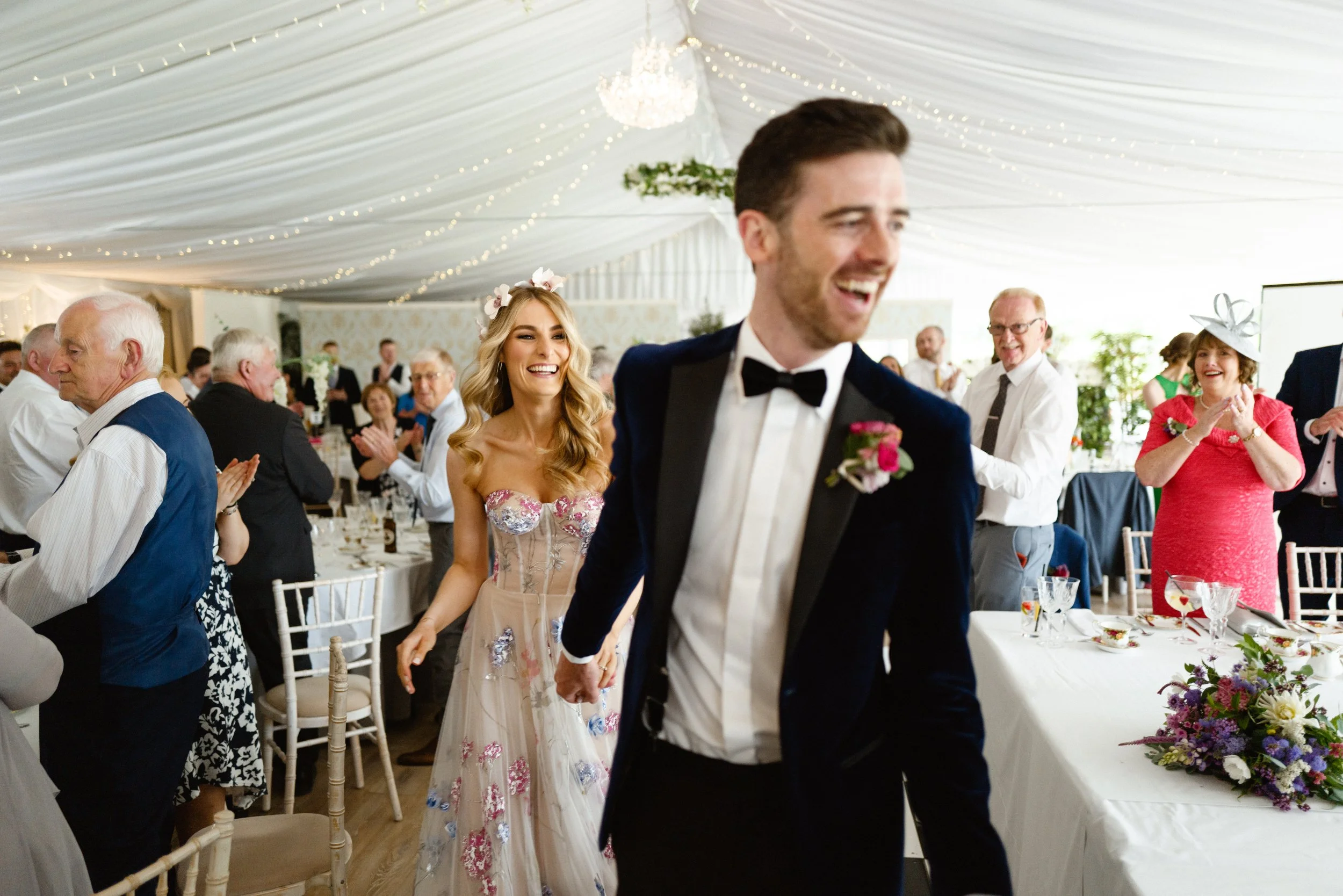 Happy wedding reception with guests clapping and smiling, bride and groom walking hand-in-hand in elegant attire, decorated venue with draped ceiling and floral arrangements.